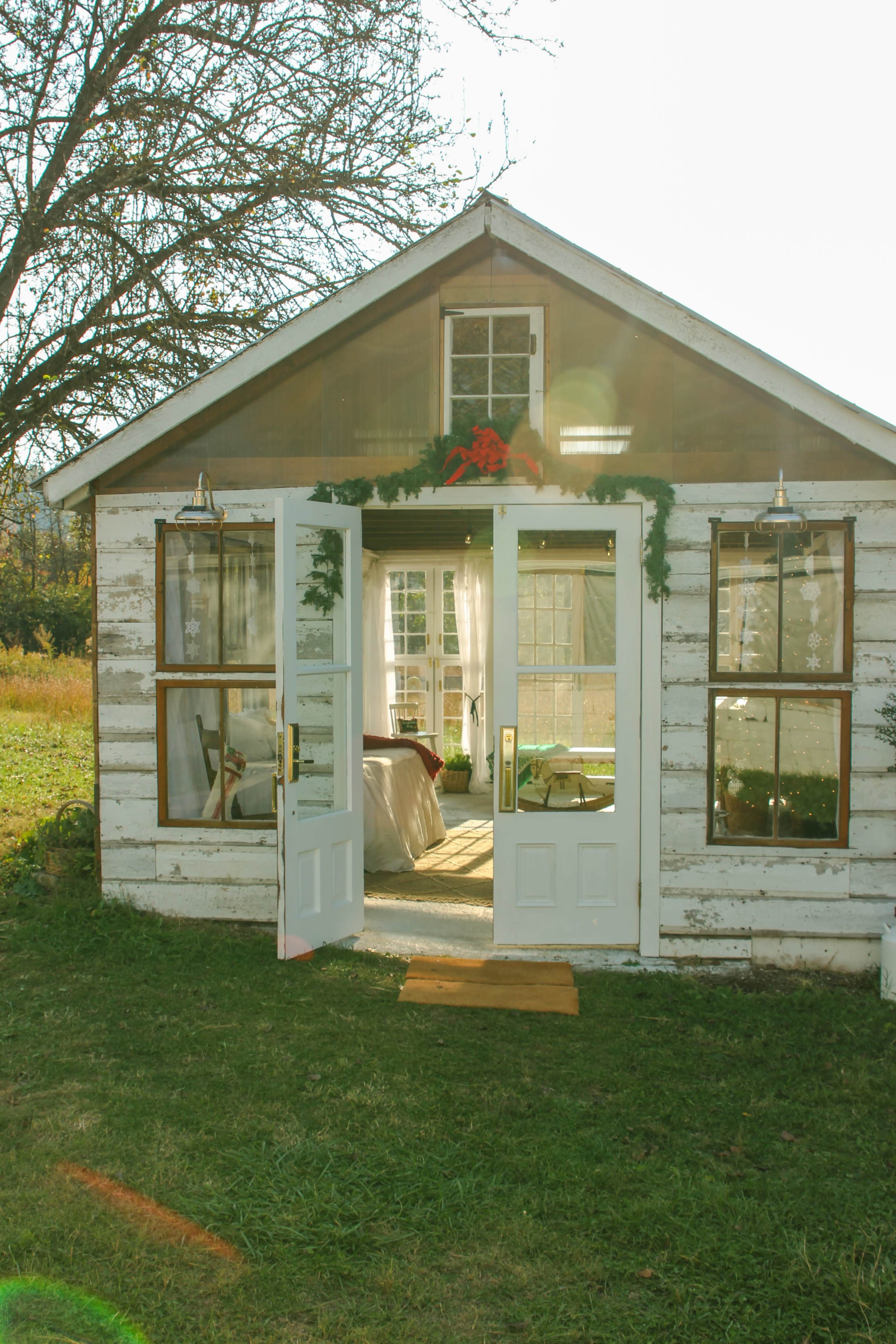 A small wooden shed with large windows and double doors opens into a sunny interior filled with plants and natural light.