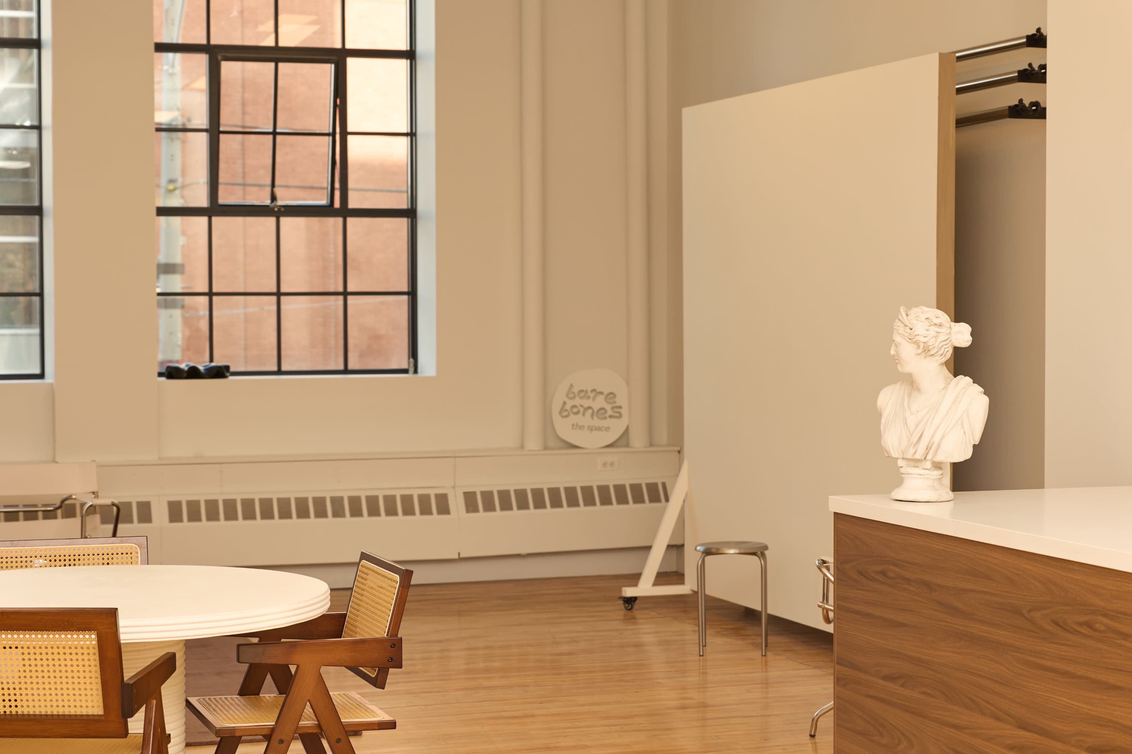 The image shows a minimalist interior space featuring a dining table with wooden chairs, a white sculpture on a countertop, and a large window providing natural light.
