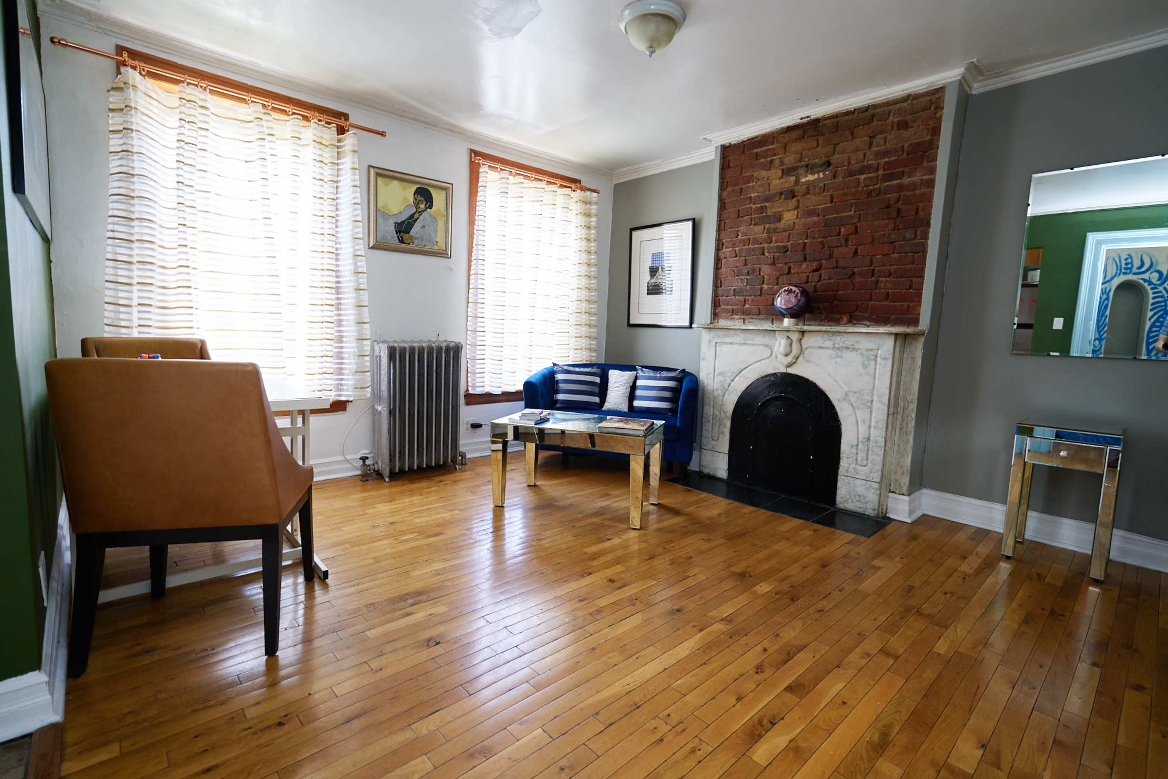 The room features wooden floors, a brick fireplace, large windows with sheer curtains, and modern furniture including a blue sofa and a glass table.