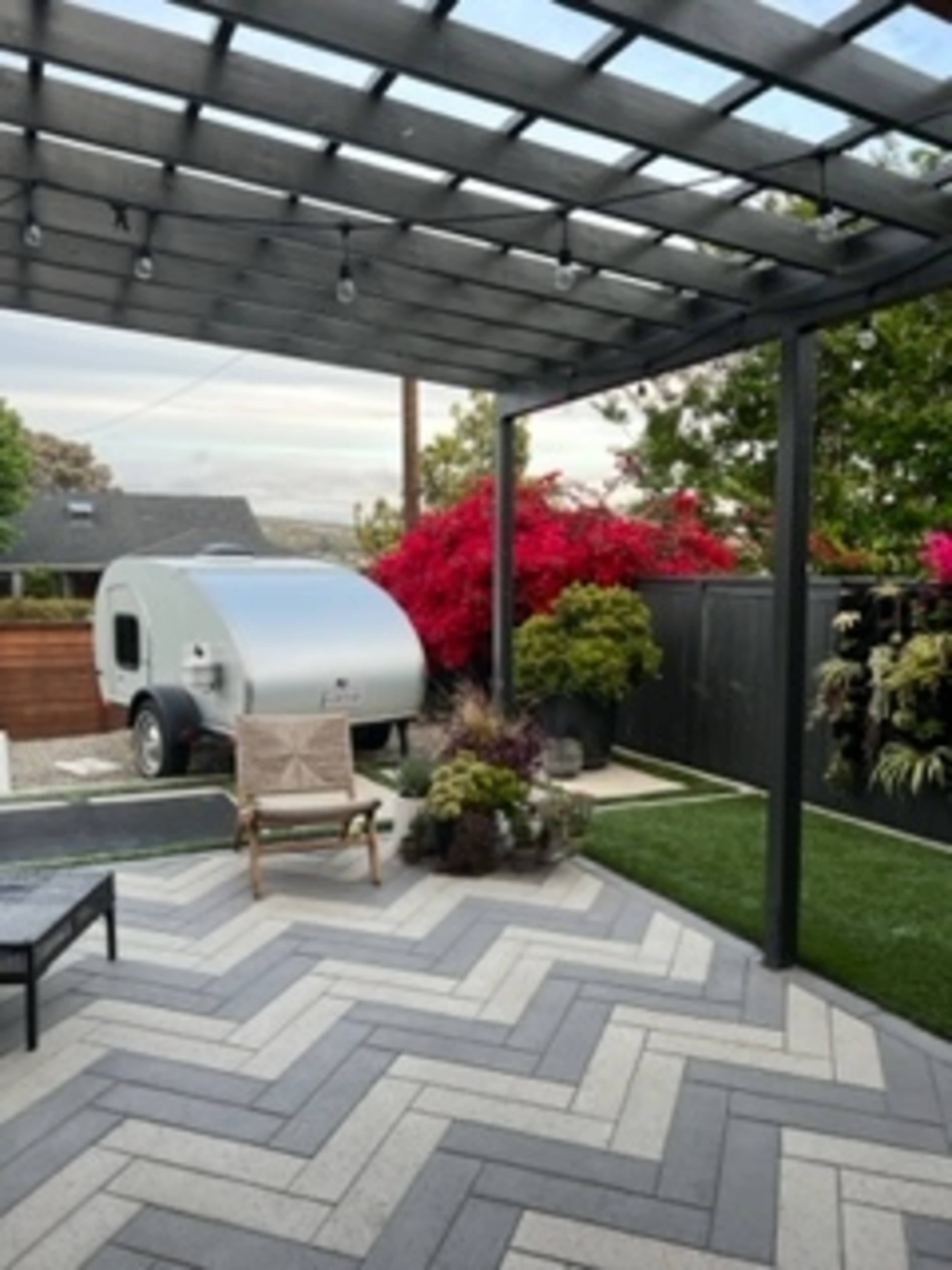 A modern outdoor patio features a pergola, a trailer parked nearby, and colorful plants against a dark fence.