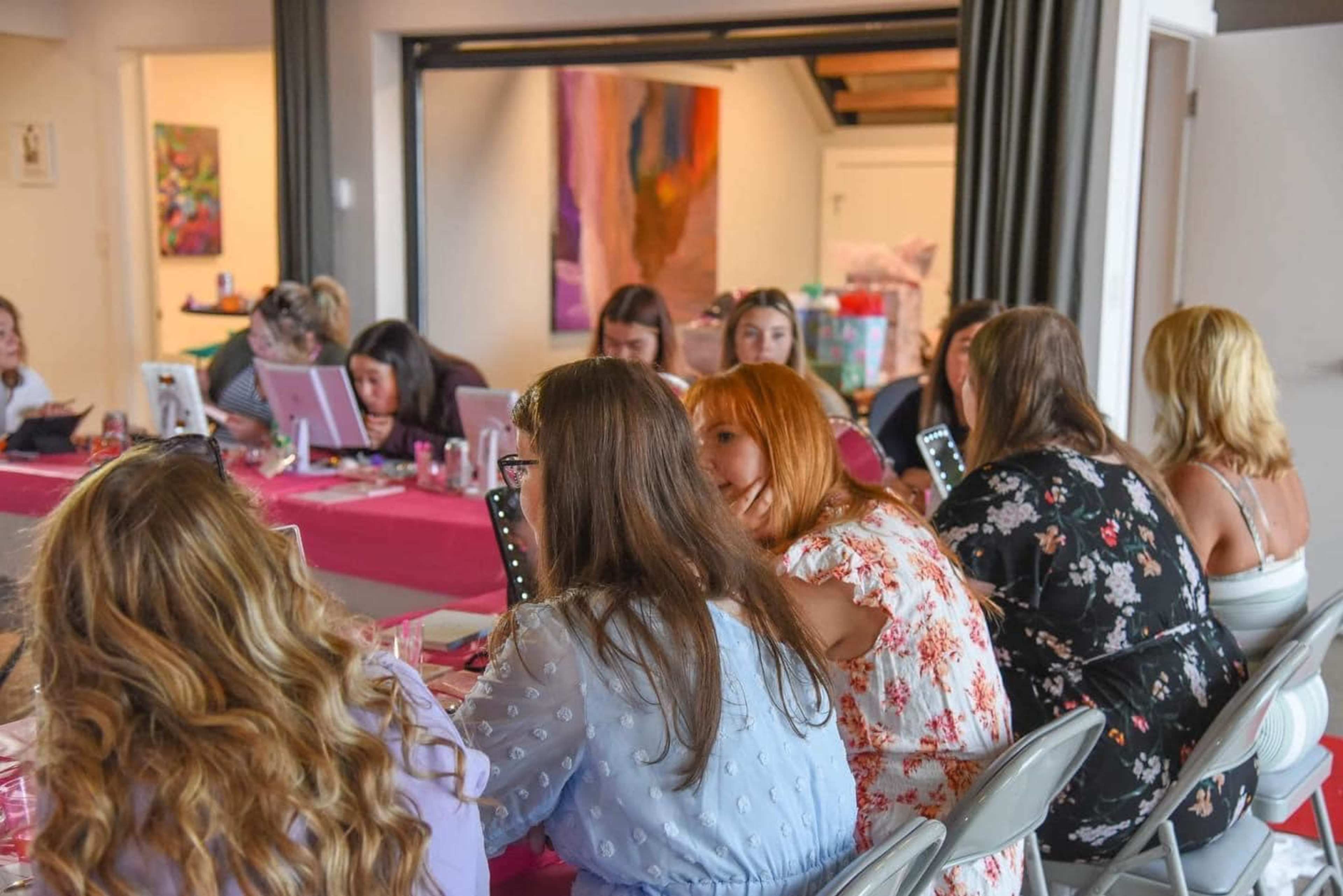 A group of women are seated at a table adorned with pink tablecloths, engaged in an activity that involves applying makeup or looking into mirrors.