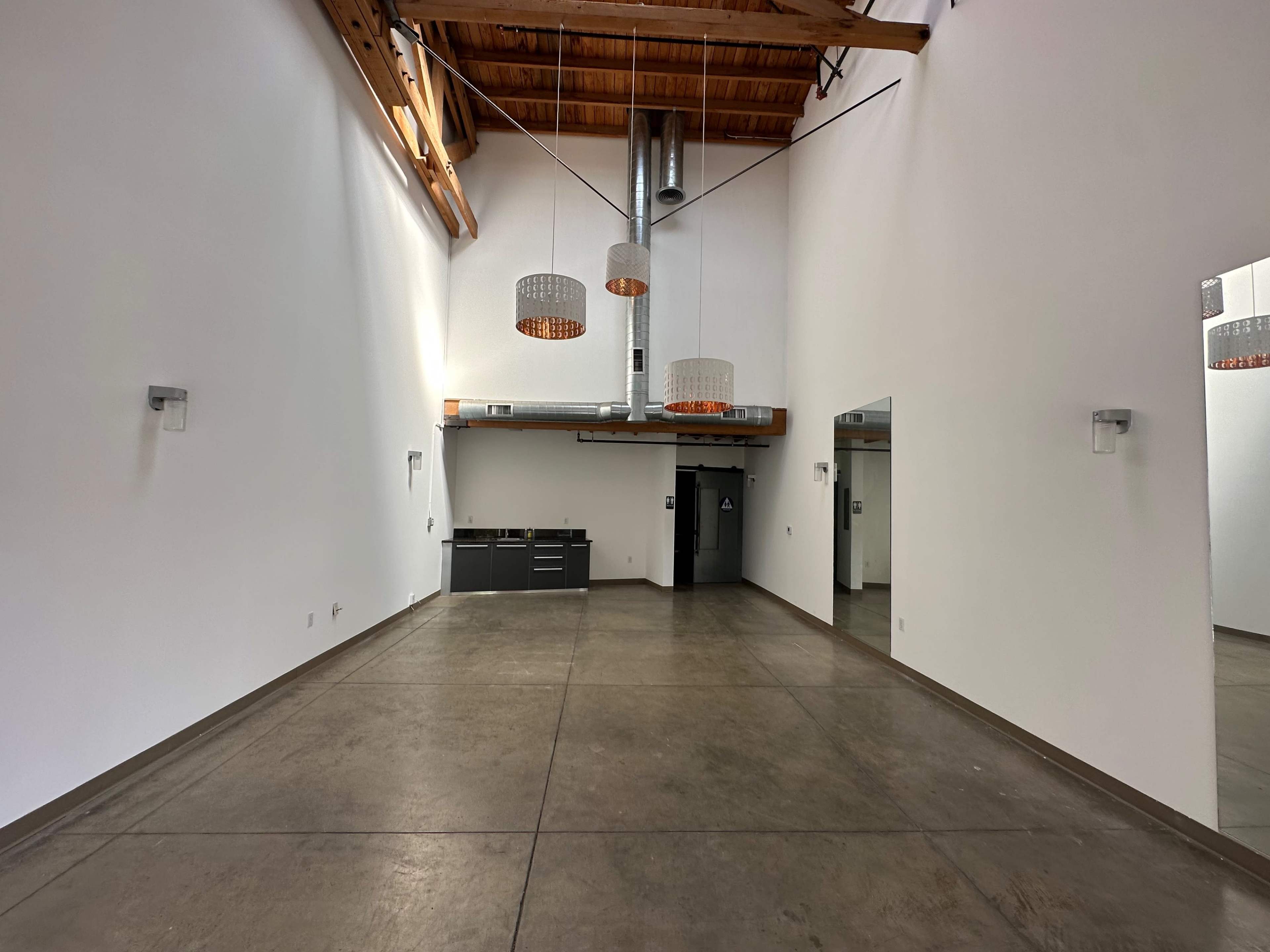 The image shows a spacious interior with high ceilings, exposed wooden beams, concrete floors, and minimalist lighting fixtures.