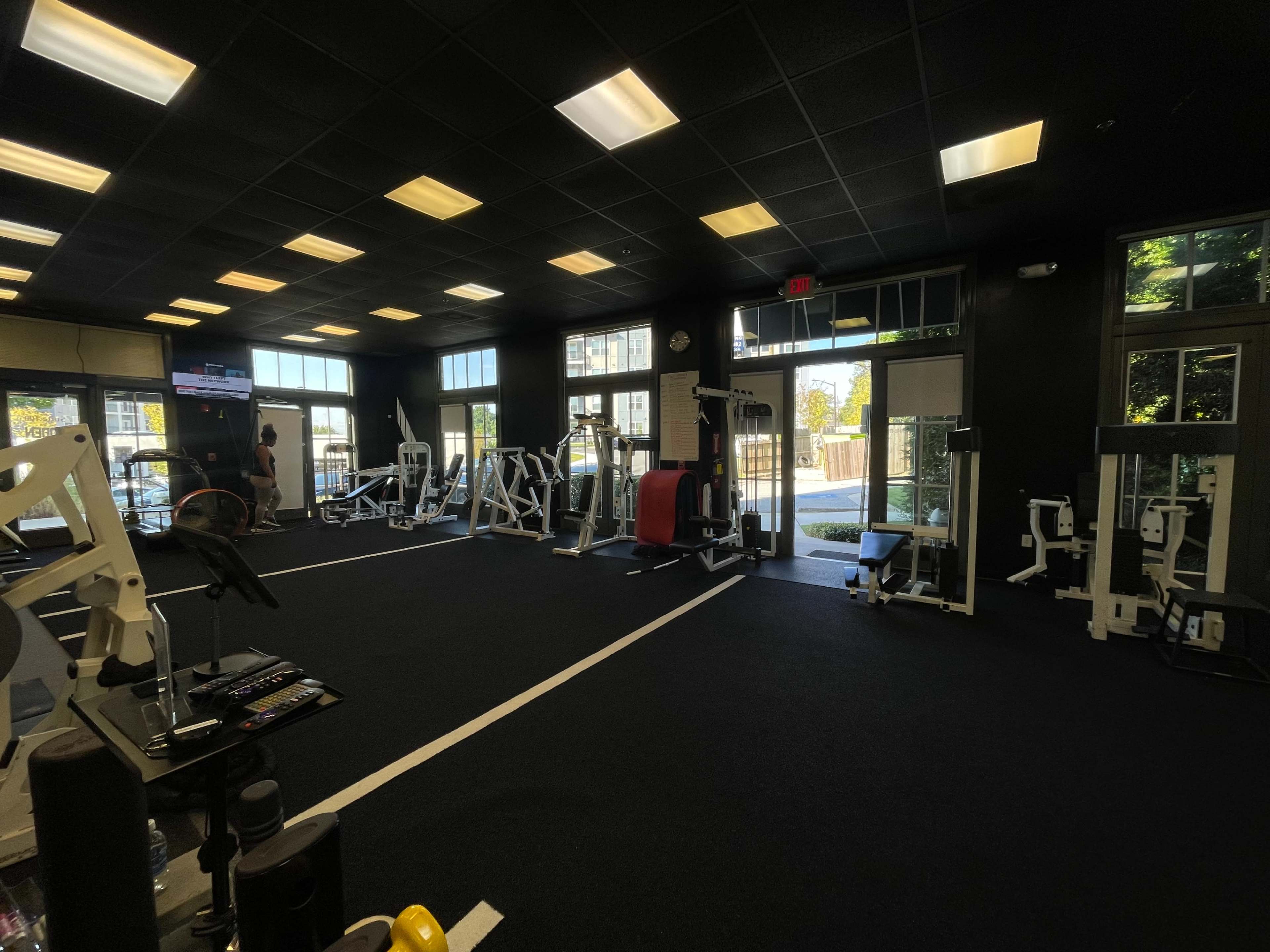 The image shows a spacious gym interior with various exercise equipment arranged throughout the room, large windows letting in natural light, and an open area near the entrance.