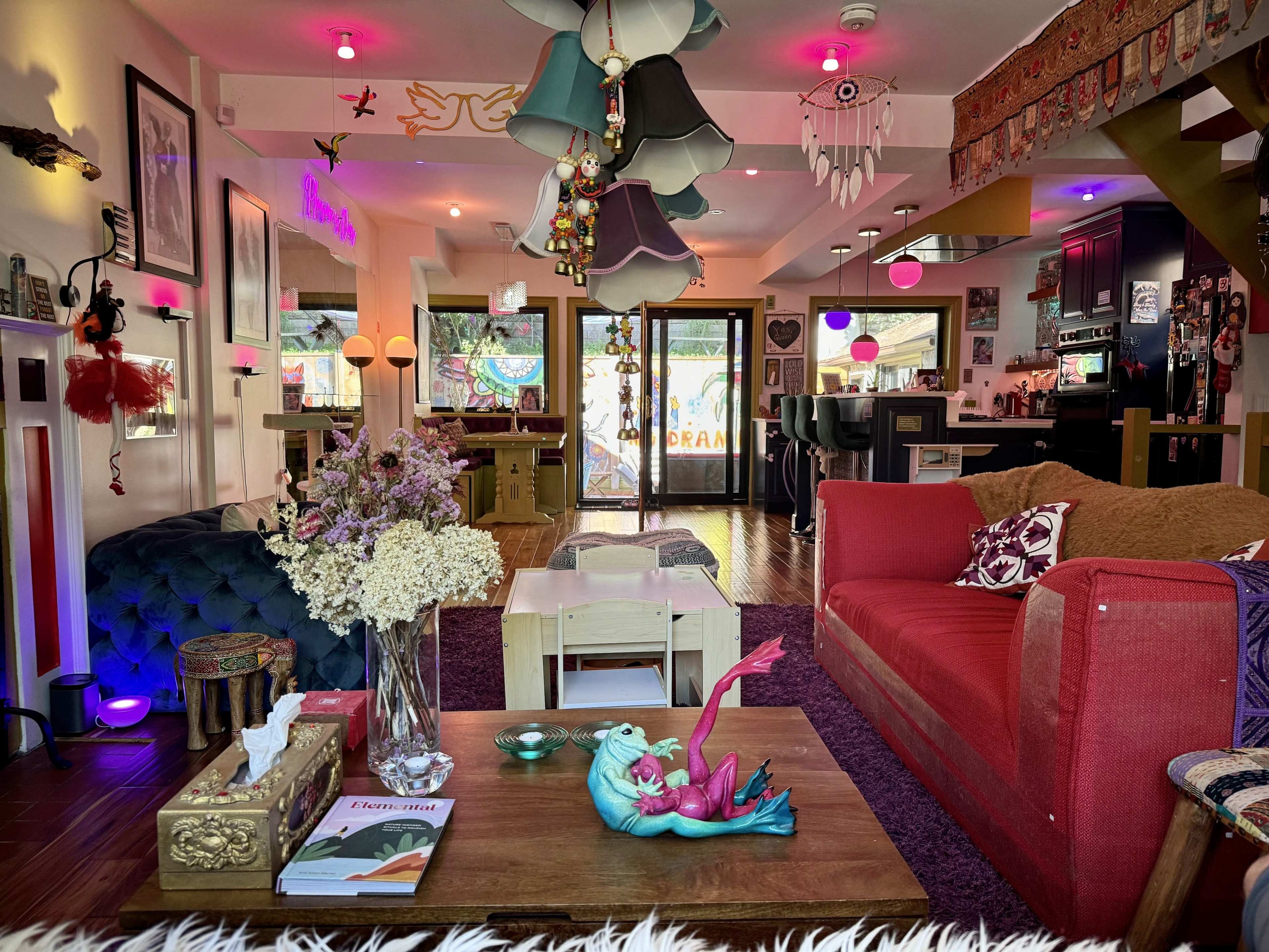 The image shows a vibrant living room filled with eclectic decor, featuring colorful lighting, a mix of furniture styles, and a decorative table with a flower arrangement and figurine.