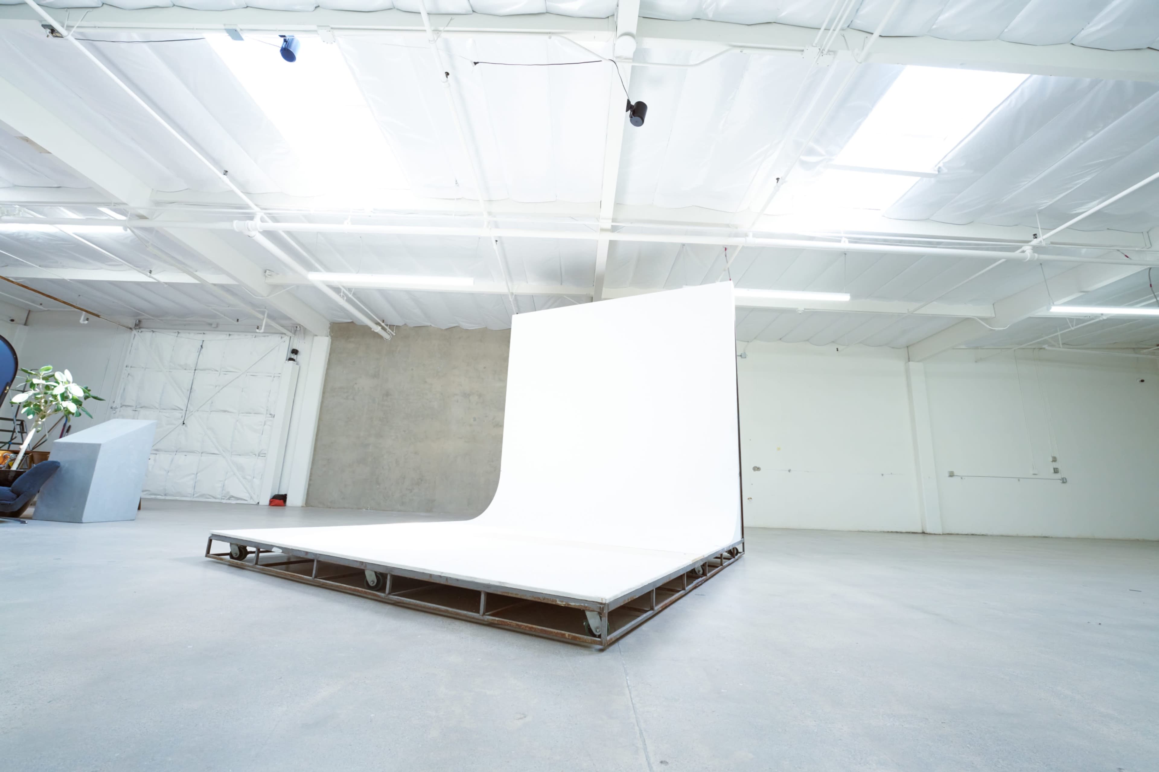 A spacious studio features a large white backdrop on a raised platform, with concrete flooring and overhead lighting.
