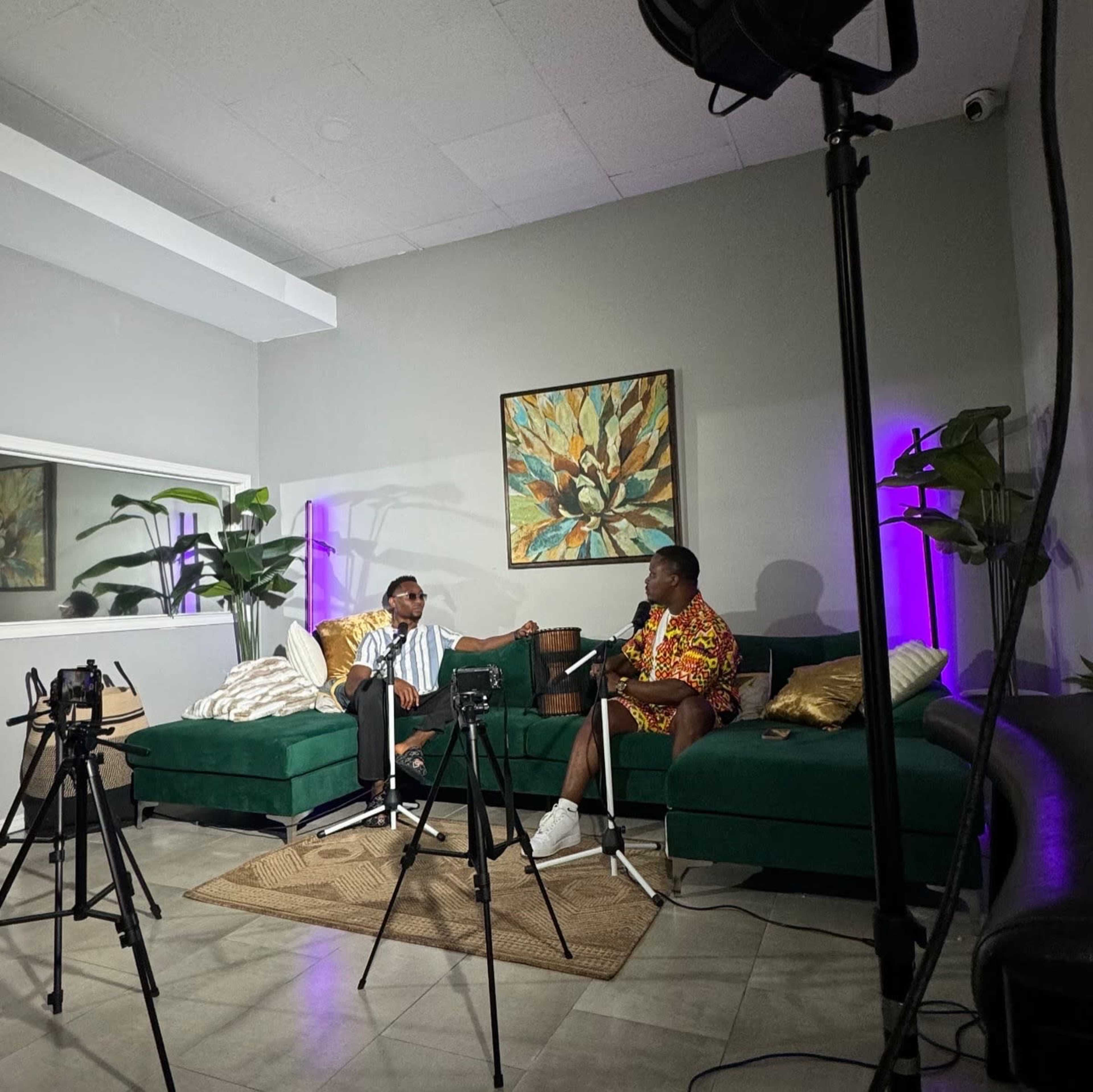 Two individuals are seated on a green sectional couch in a modern indoor setting, surrounded by plants, while being recorded by several cameras for a discussion or interview.
