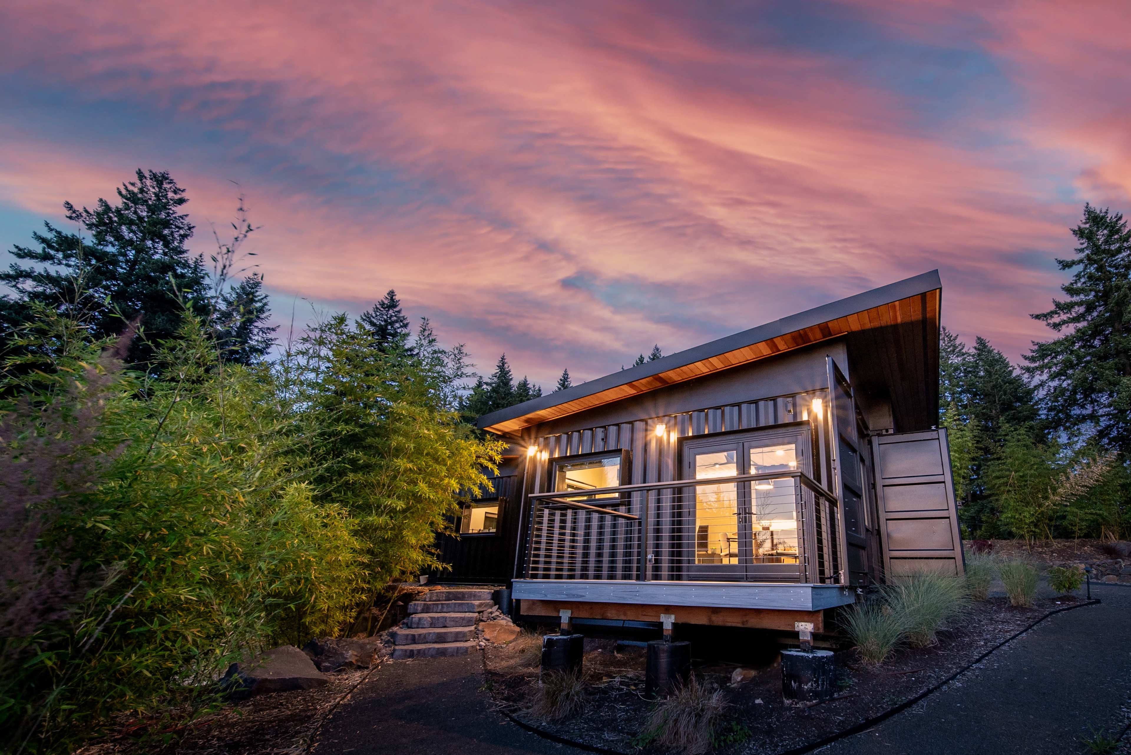 A modern cabin with large windows and a deck is illuminated under a colorful sunset sky, surrounded by trees and landscaped greenery.