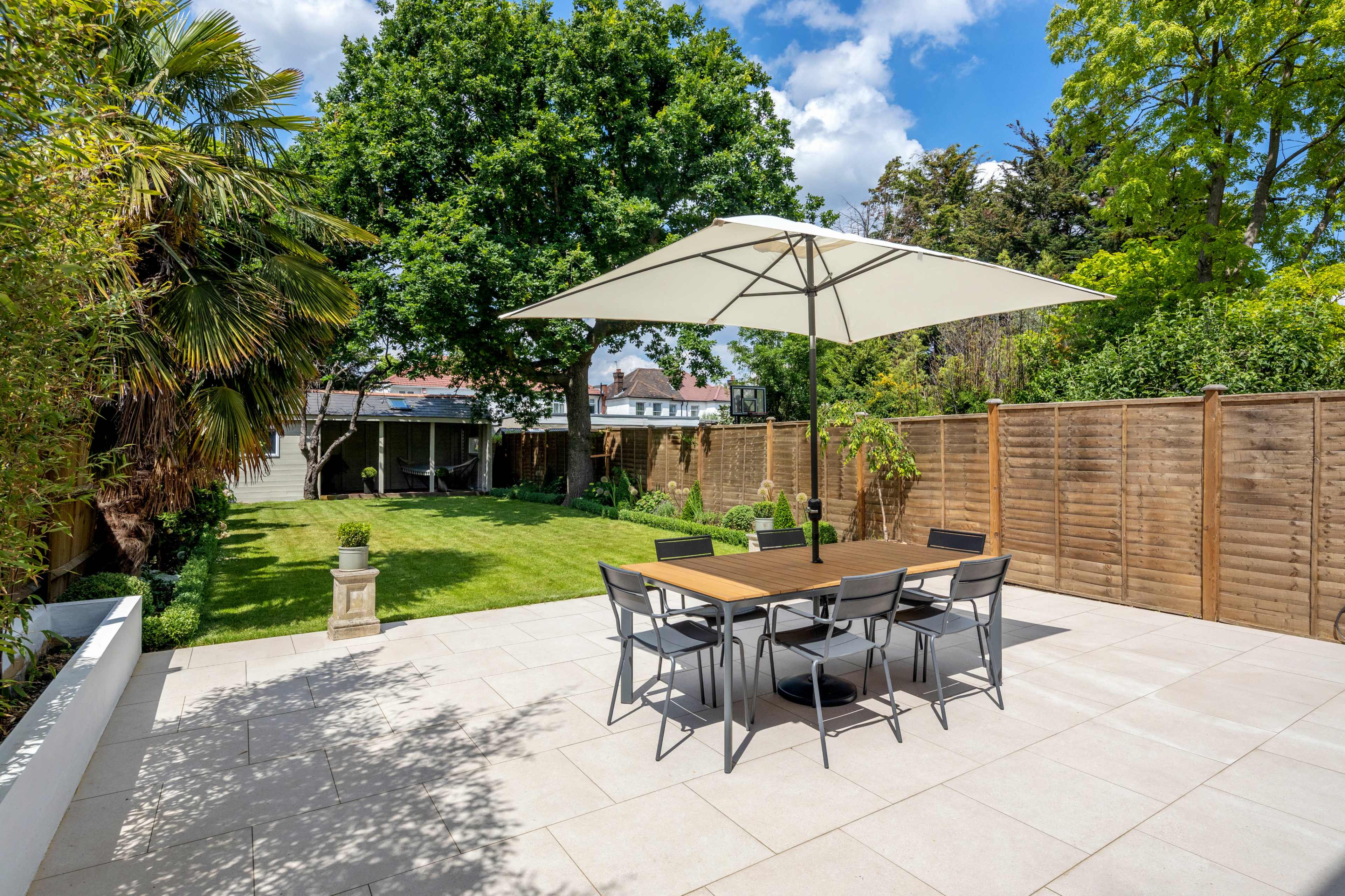 A rectangular patio with a large umbrella shades a dining table surrounded by chairs, overlooking a green lawn and trees.