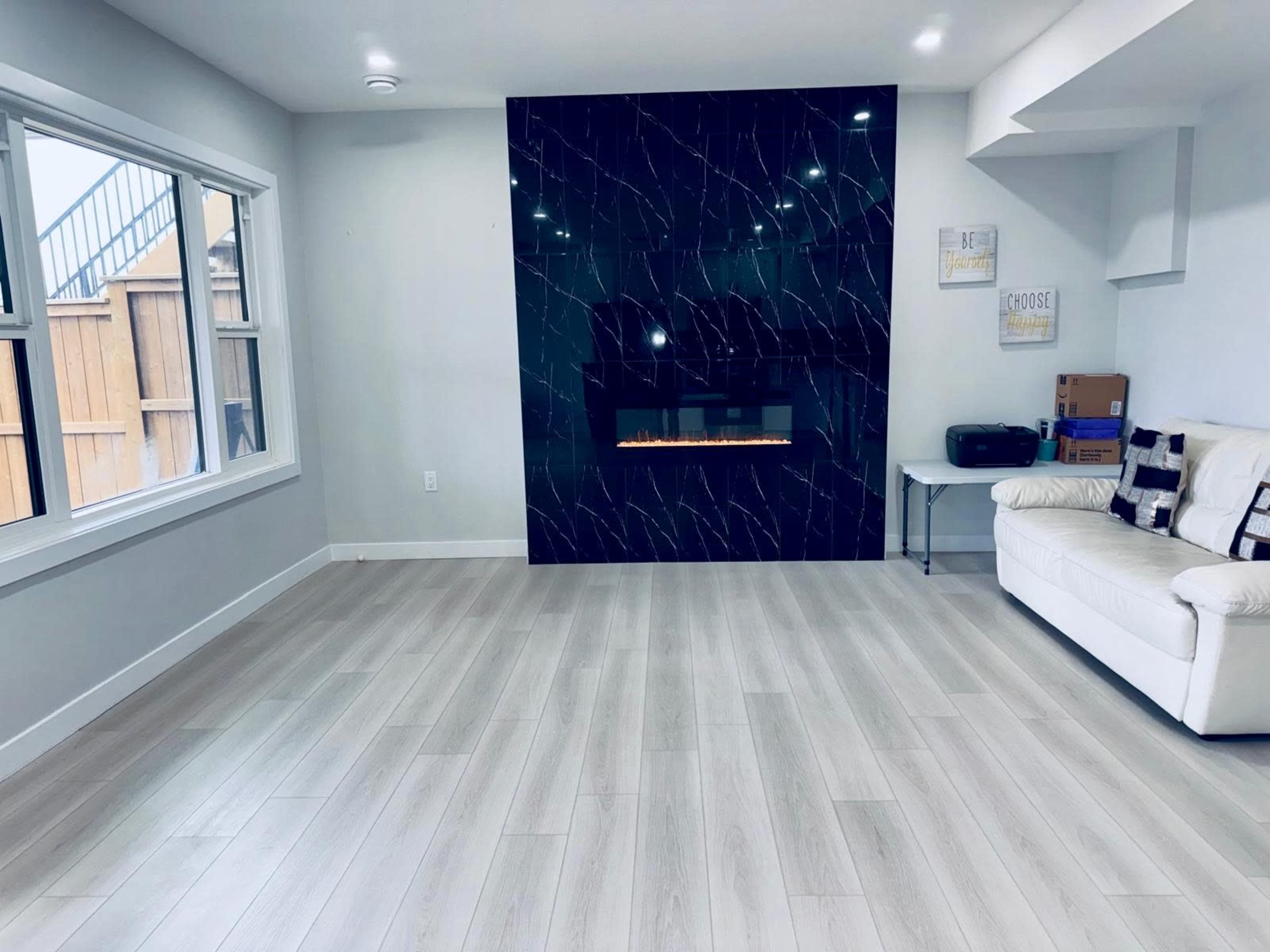 The image shows a modern living room featuring a white sofa, a sleek black fireplace wall, and large windows that allow natural light to enter.