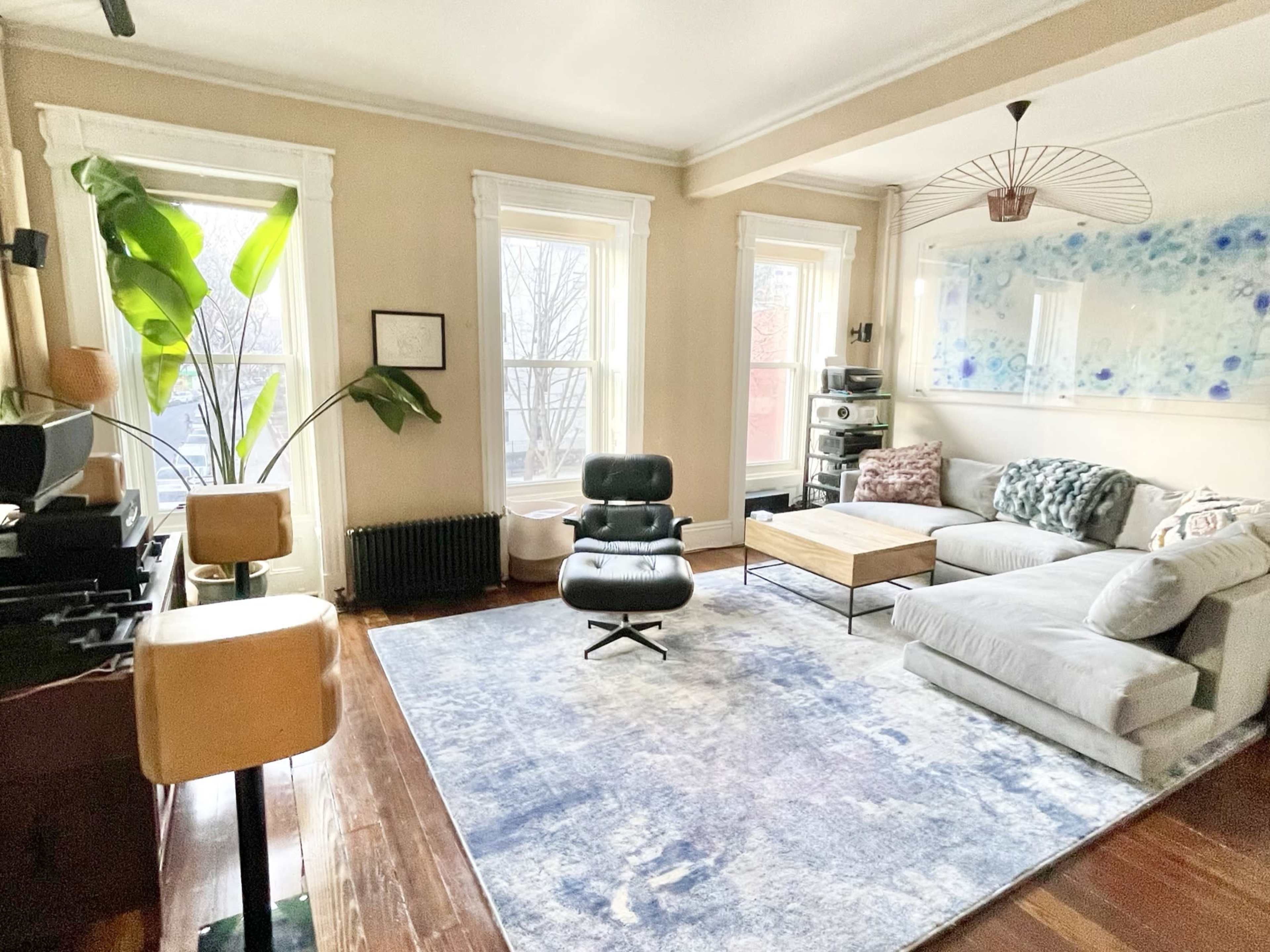 A cozy living room with large windows, a sectional sofa, a recliner chair, a coffee table, and a rug on hardwood flooring.