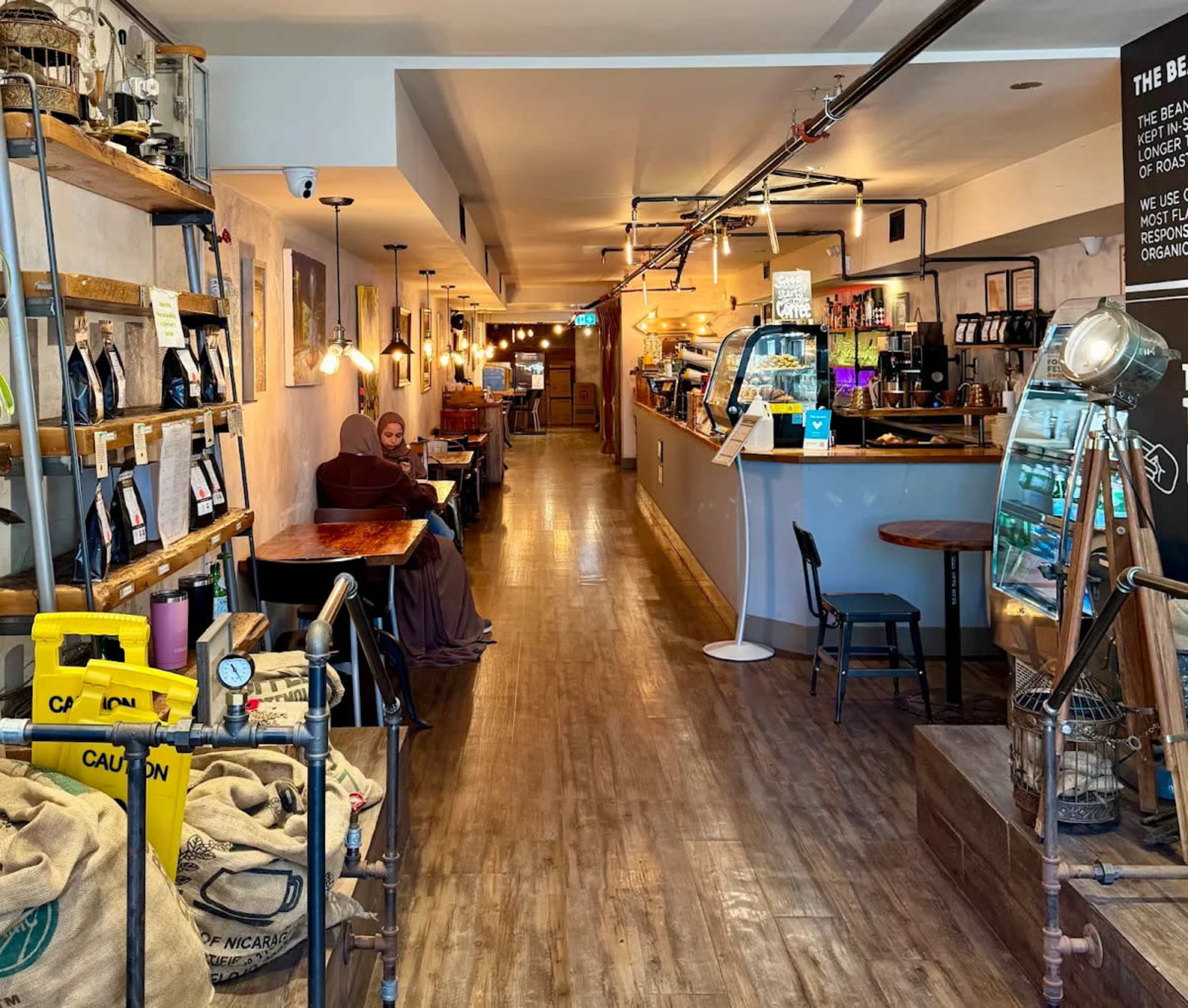The image shows a cozy café interior with wooden flooring, shelves displaying various products, and a service counter at the end.