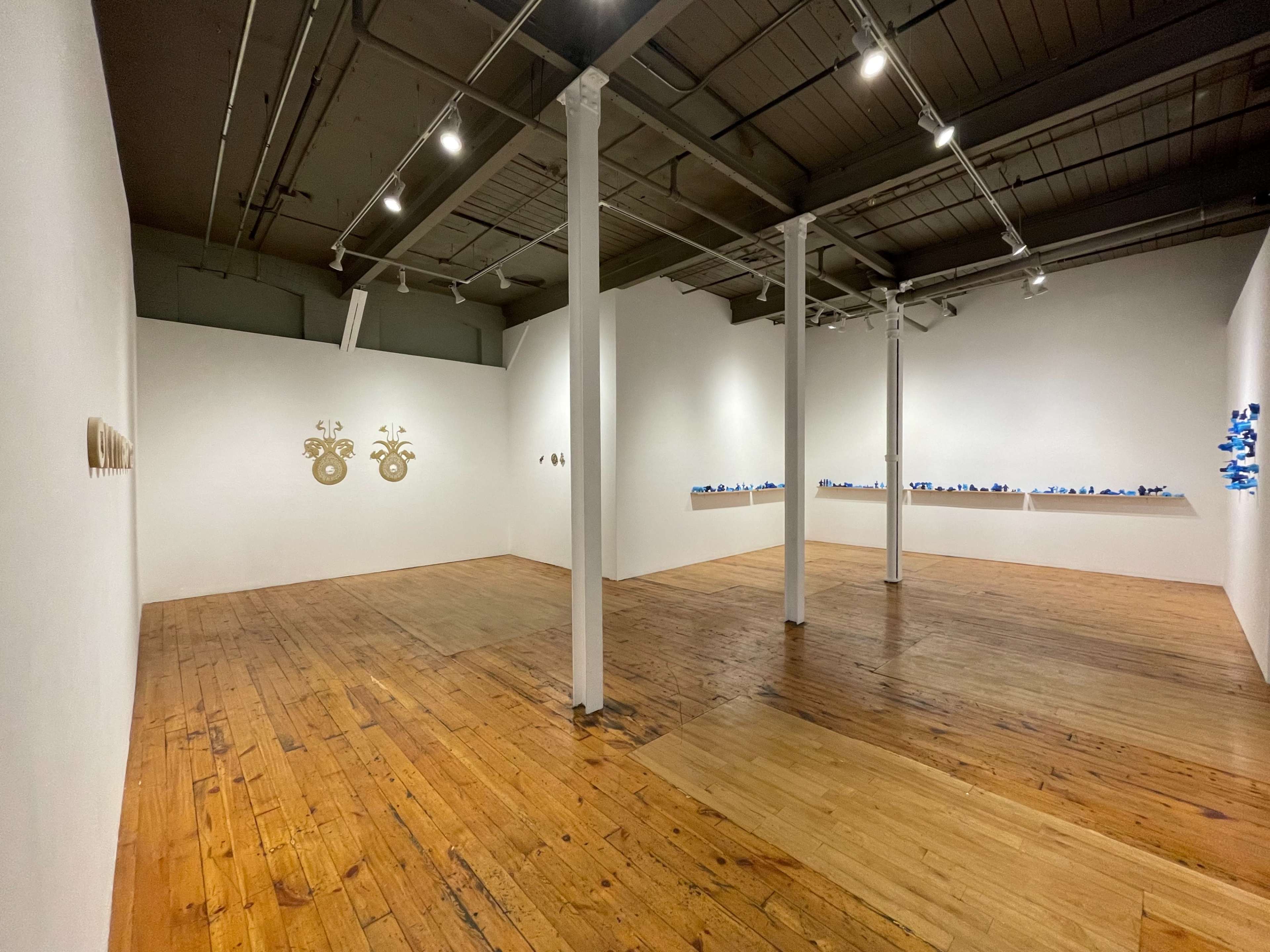 A spacious gallery with wooden flooring and white walls displaying various artworks, including two gold circular pieces on the left and a line of blue items on the right.