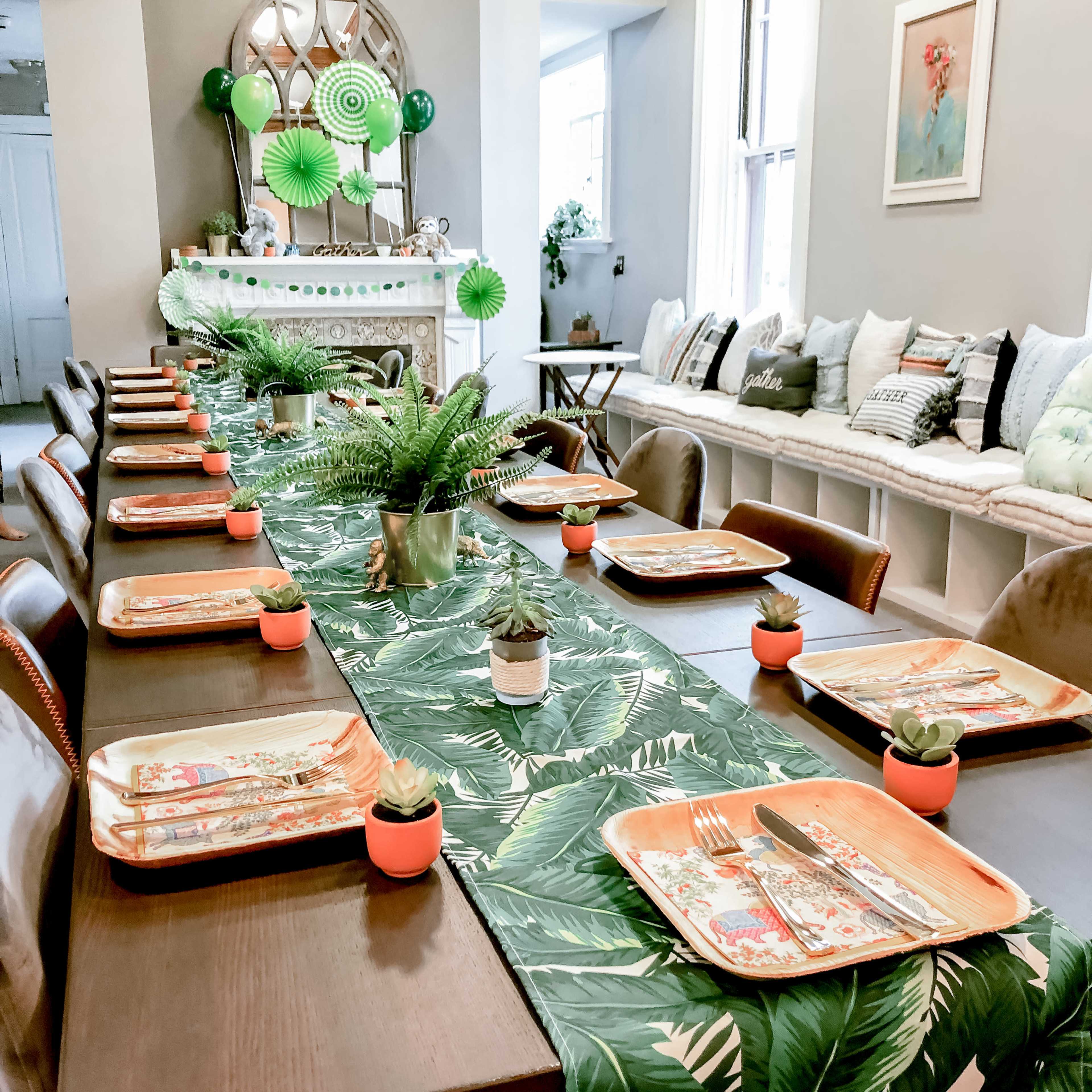 A long dining table is set with tropical-themed tableware, surrounded by chairs, and decorated with greenery and small potted plants.