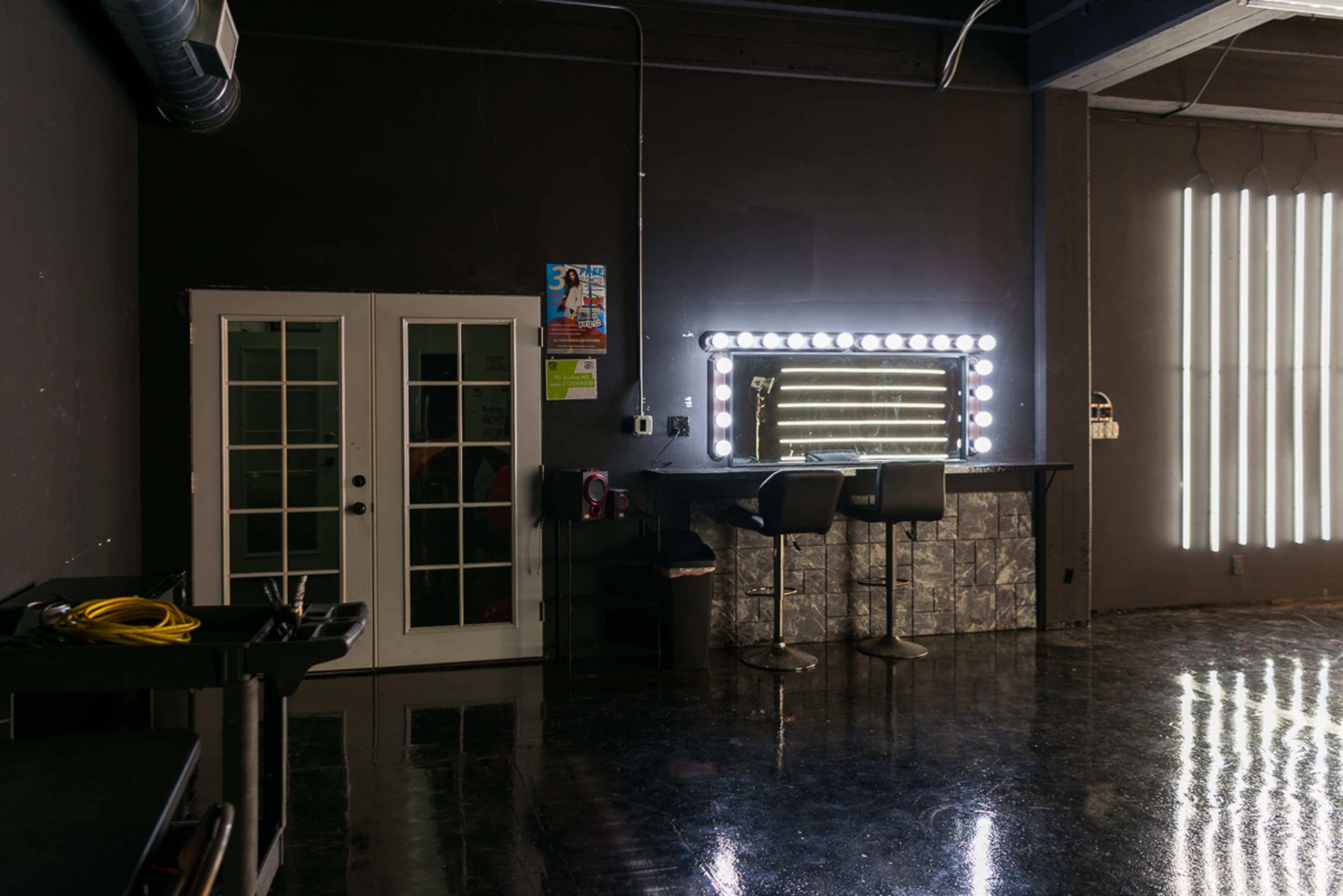 The image shows a dimly lit room with a mirror surrounded by bright lights, two bar stools, and a glass double door leading to another space.