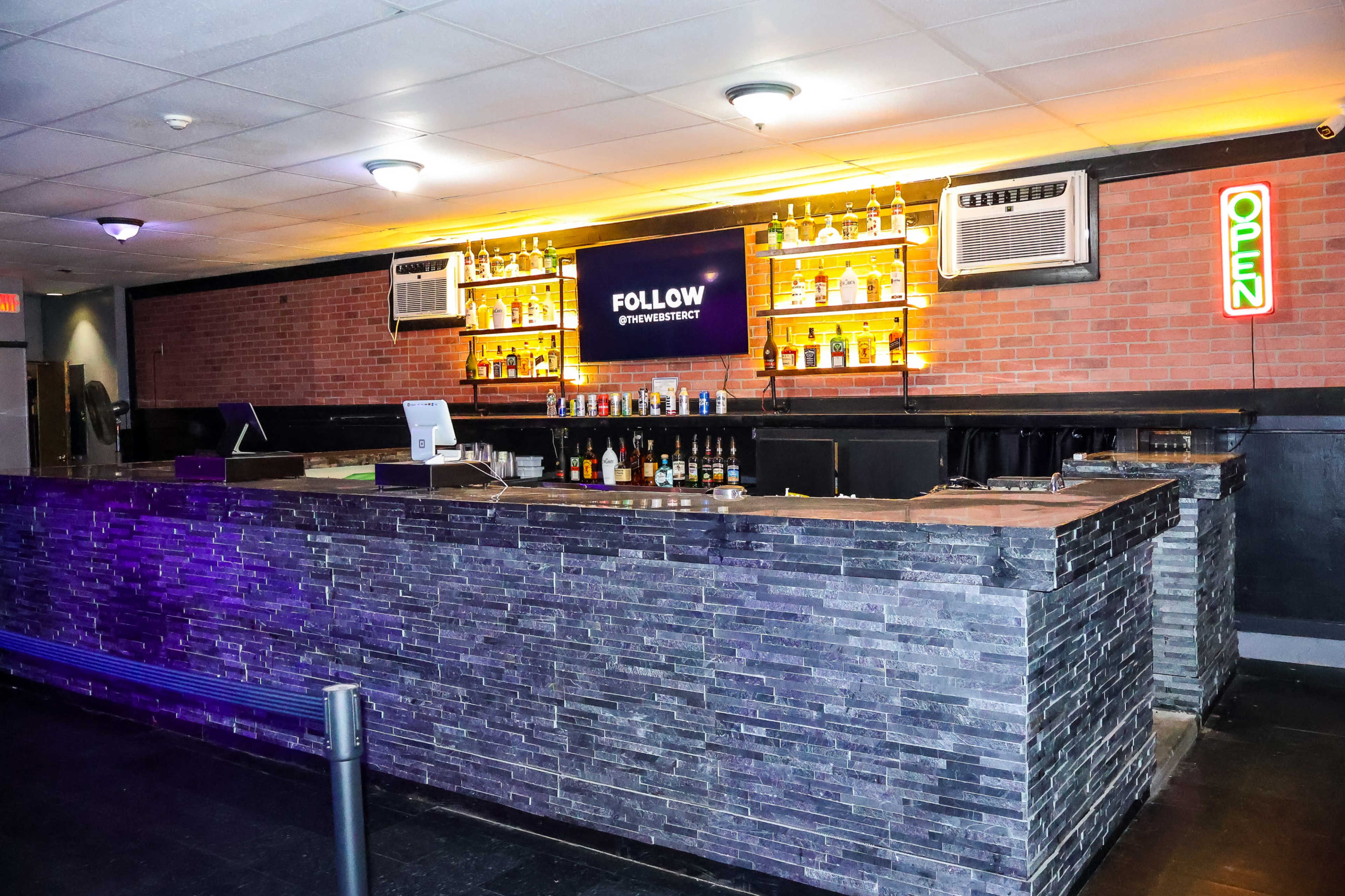 A bar with a stone counter, illuminated shelves of liquor bottles, and a neon "OPEN" sign.