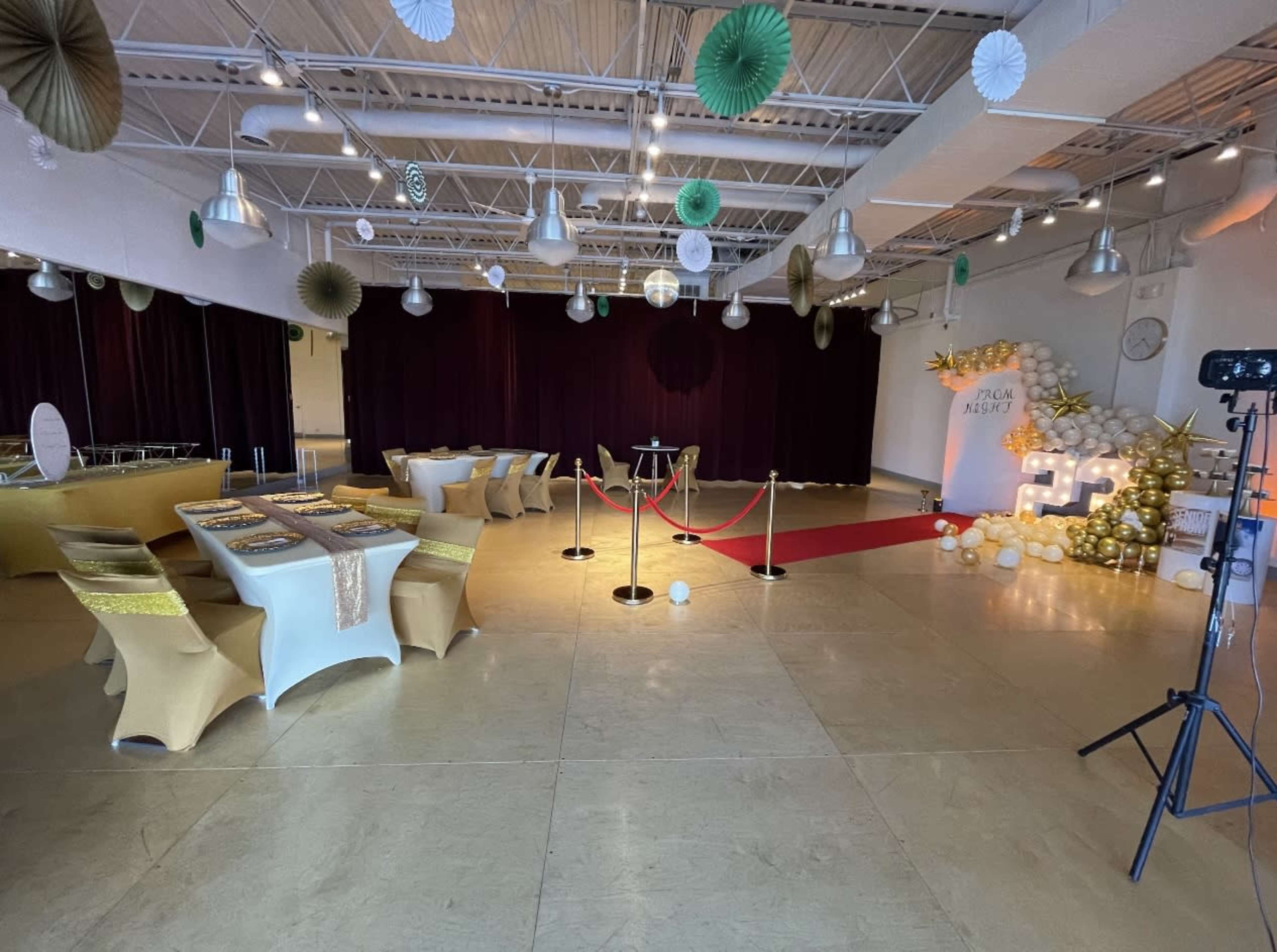 The image shows a decorated event space featuring tables set with gold and white tablecloths, a red carpet area with stanchions, and a backdrop for photographs.