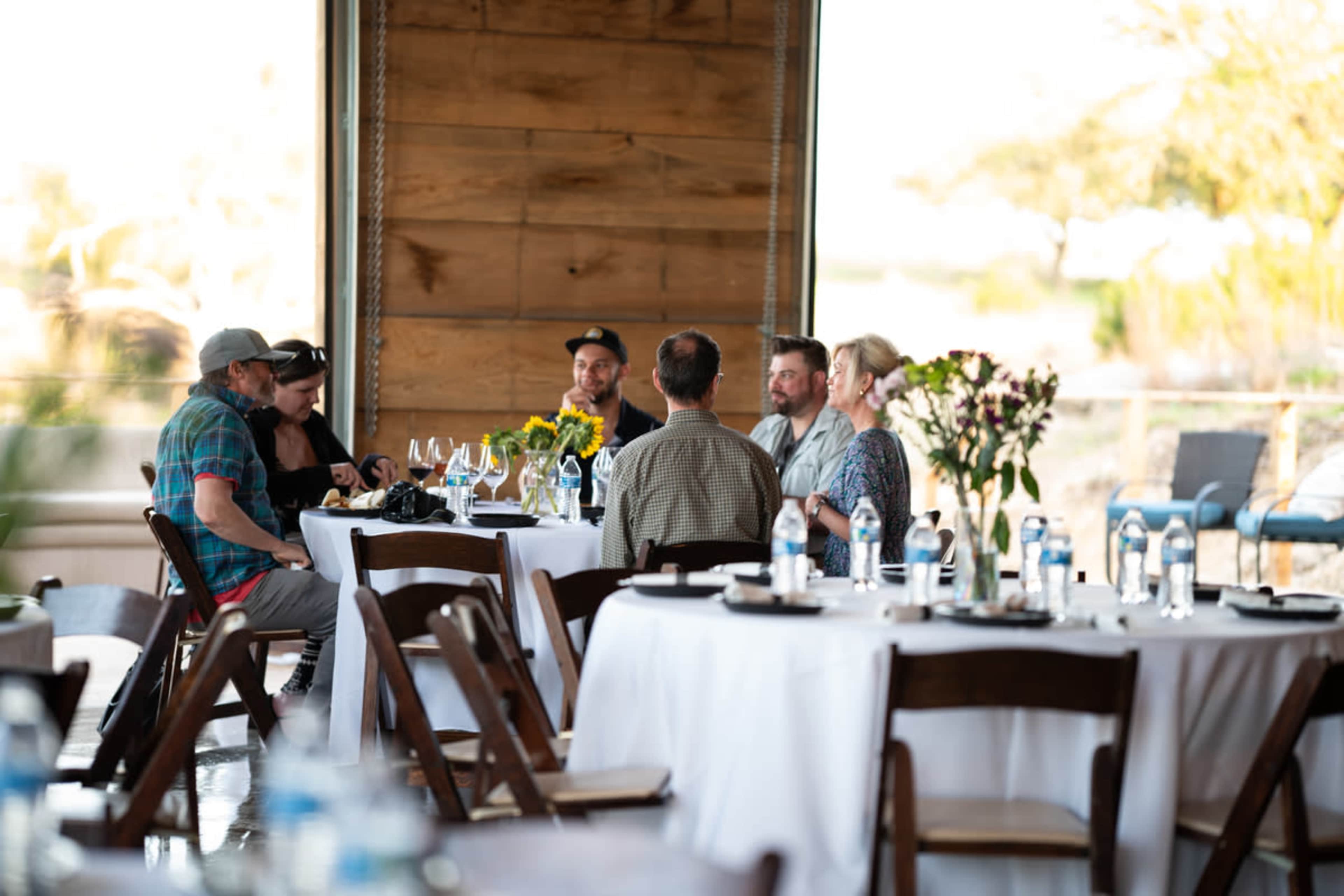 A group of six people is seated around a table in a well-lit venue, with water bottles and floral arrangements on the tables.