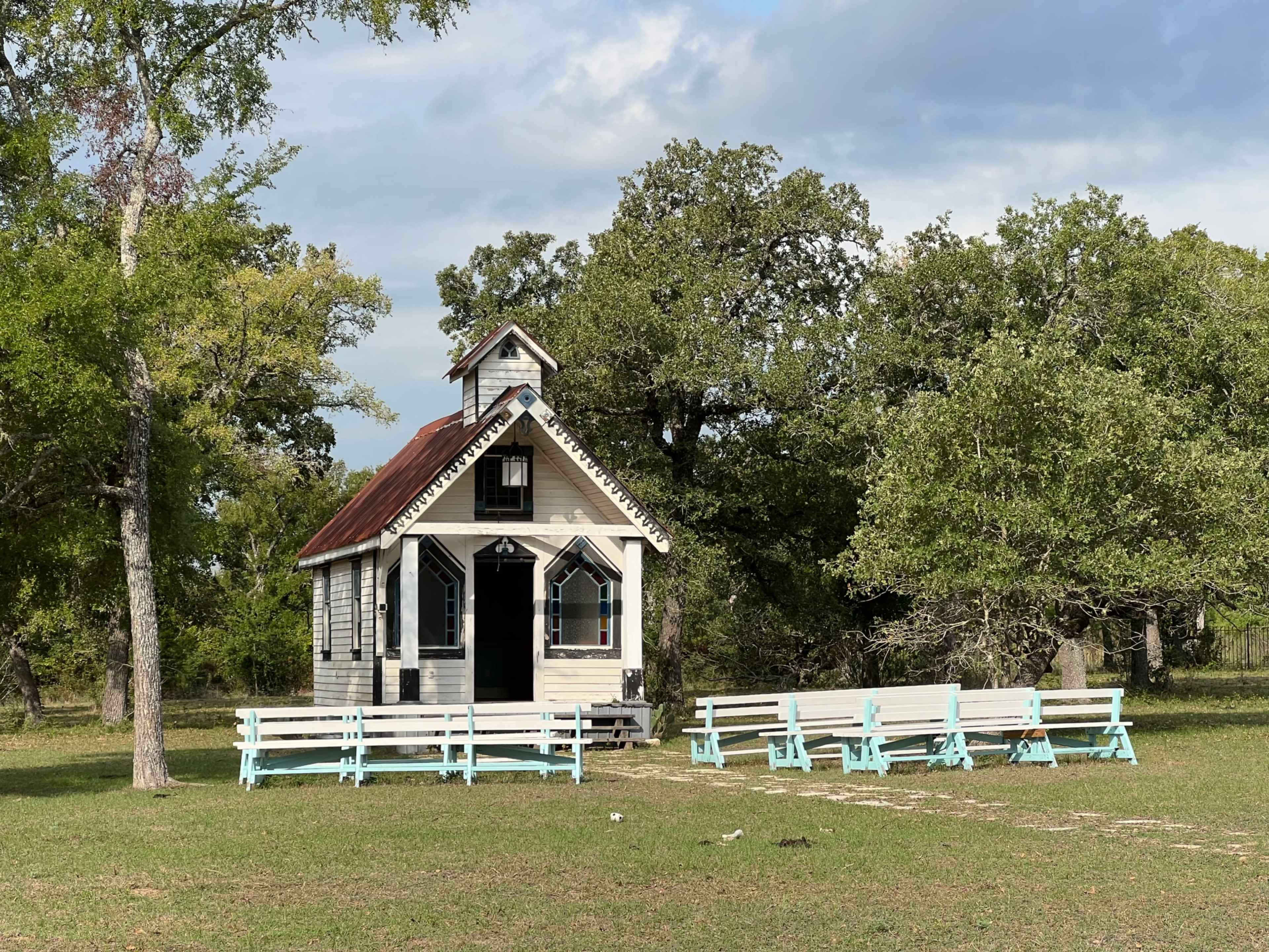 A small, white wooden chapel with a red roof sits among green trees, surrounded by several light blue benches arranged in front of it.