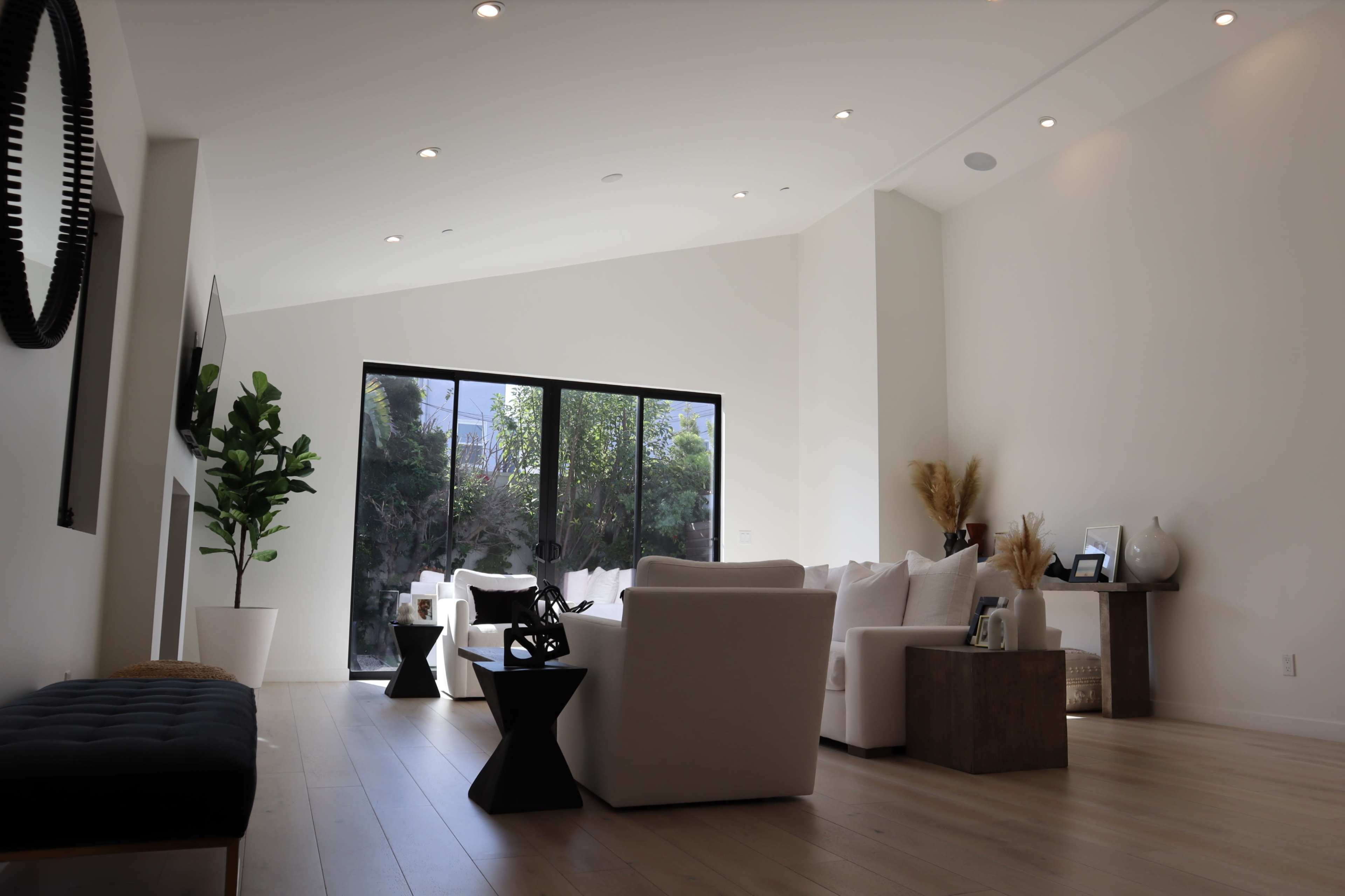 A modern living room with white sofas, a black side table, a large window overlooking greenery, and a small plant in a pot.