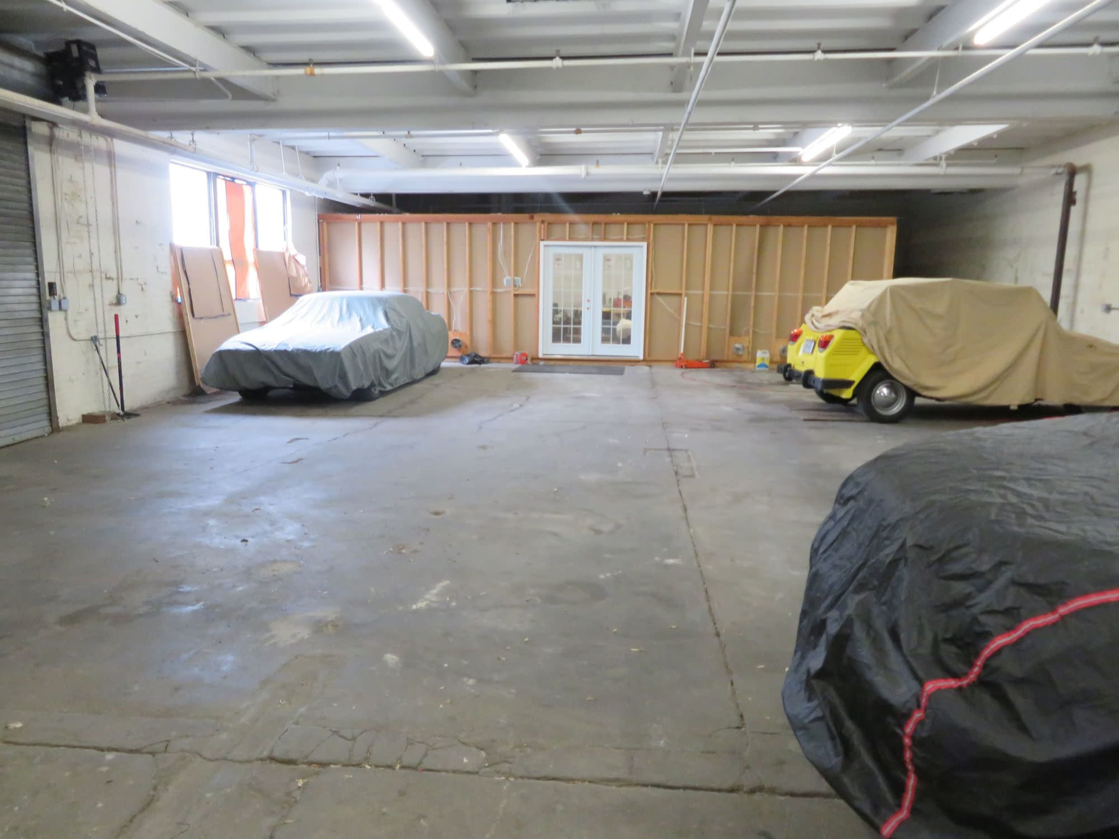 The image shows a spacious garage with several covered vehicles, a small enclosed area at the back, and natural light coming through windows.