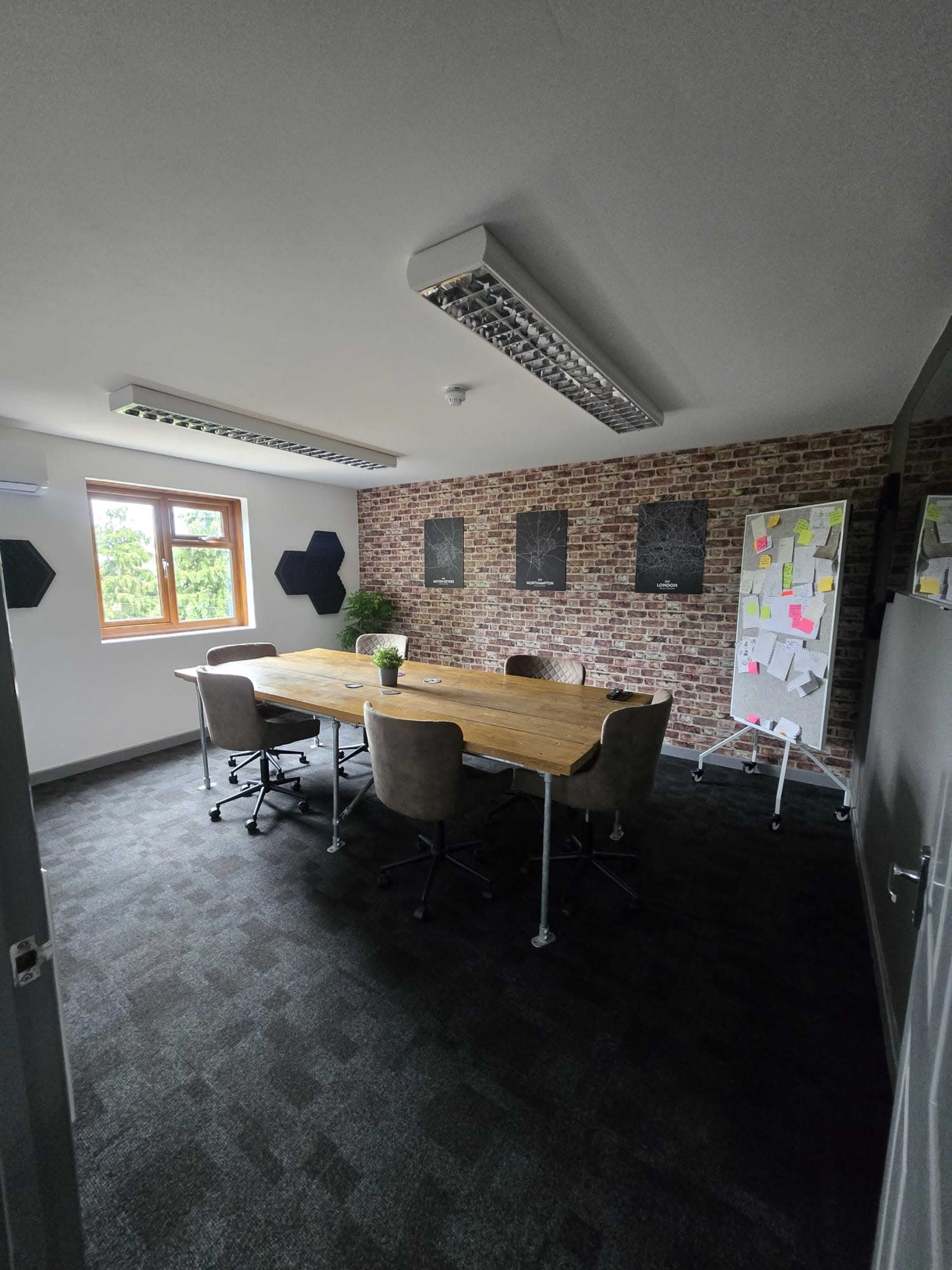 The image shows a conference room with a wooden table, five chairs, a large window, a plant, and a whiteboard covered in notes on a brick wall.