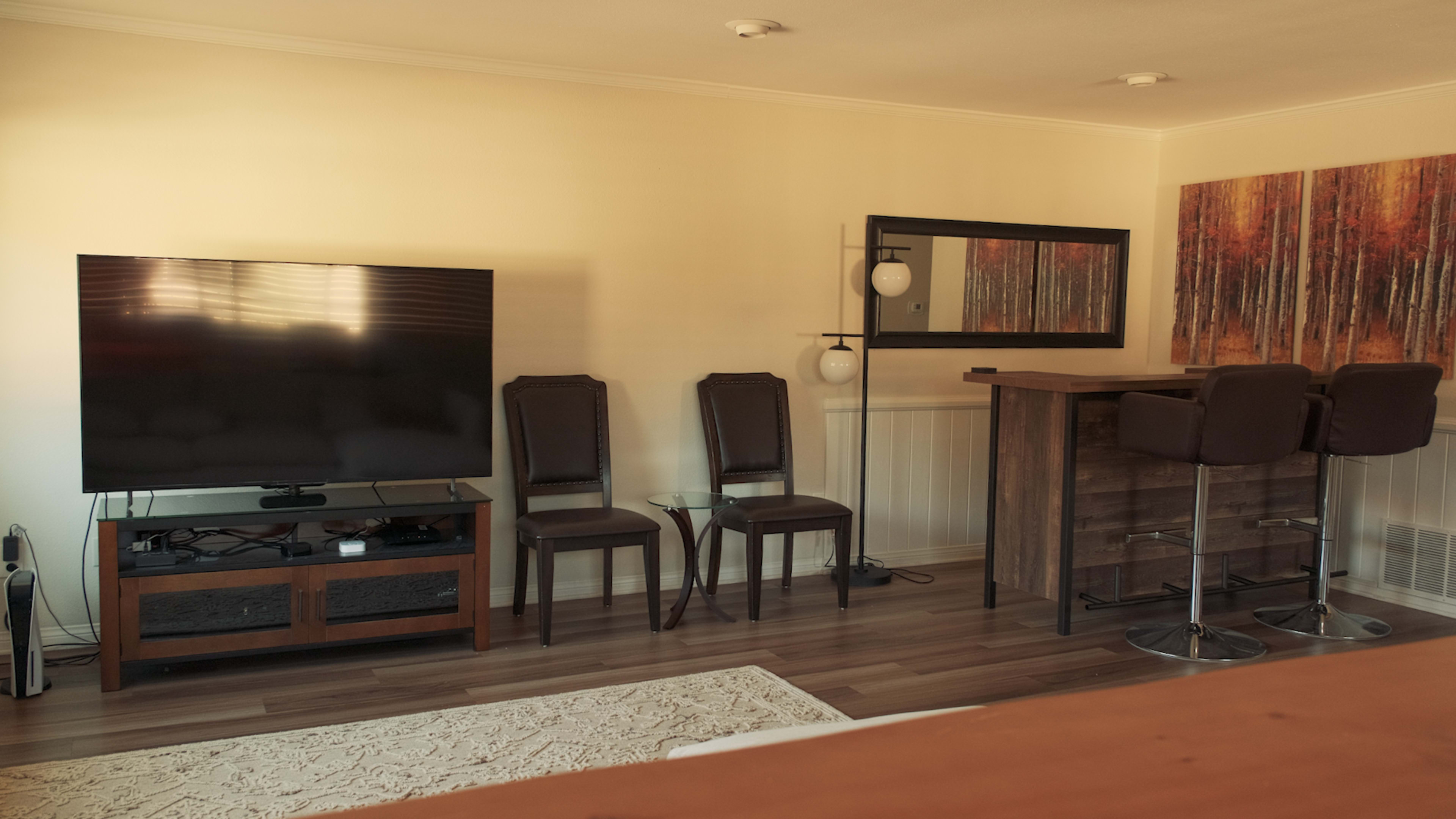 A living room features a large television, two chairs, a bar with stools, and a decorative painting of trees on the wall.