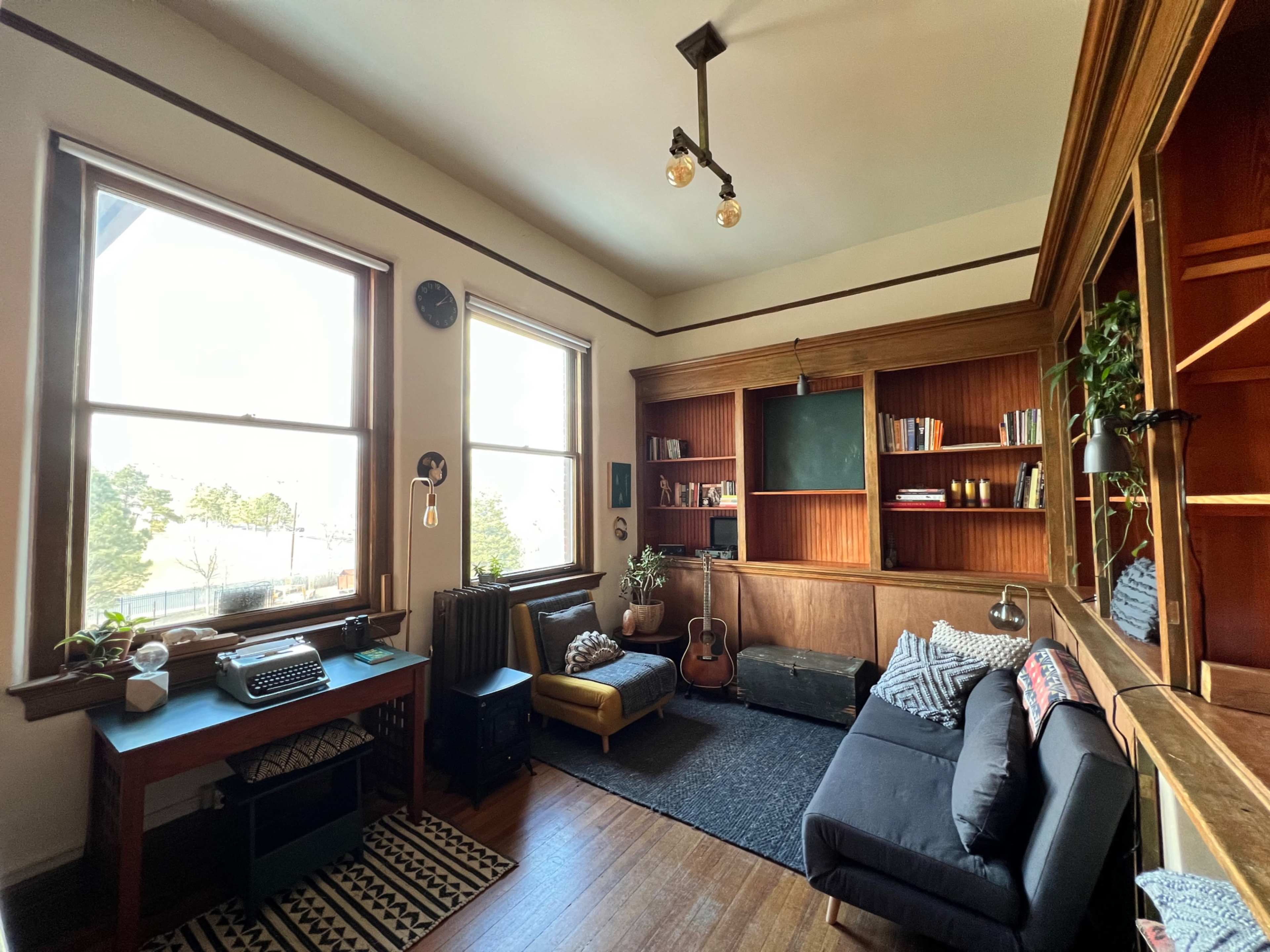 A cozy room featuring large windows, a wooden bookshelf, a black sofa, a vintage typewriter on a desk, and a guitar leaning against the wall.