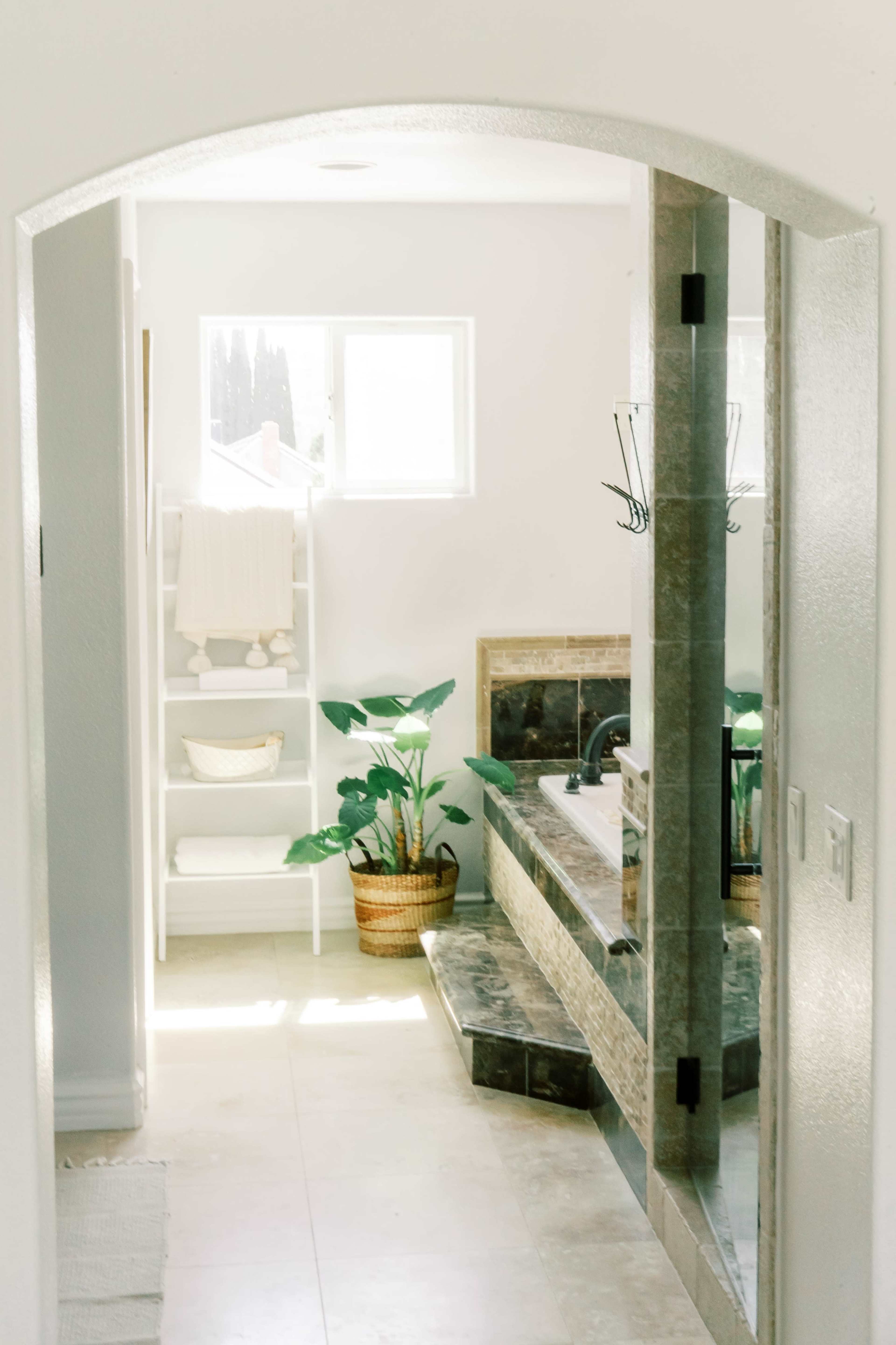 The image shows a bright, modern bathroom with a large tub, potted plants, and a tall shelving unit against a white wall.