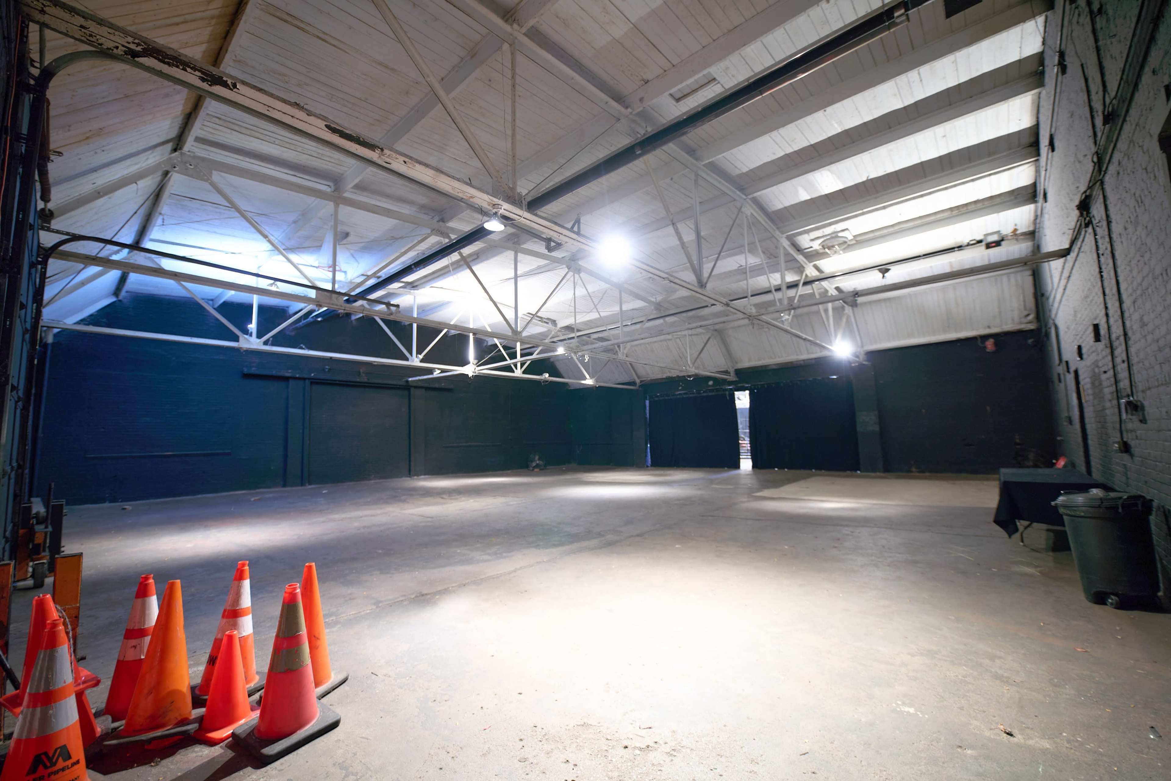 The image shows a large, empty warehouse space with high ceilings, several orange traffic cones in the foreground, and minimal lighting overhead.