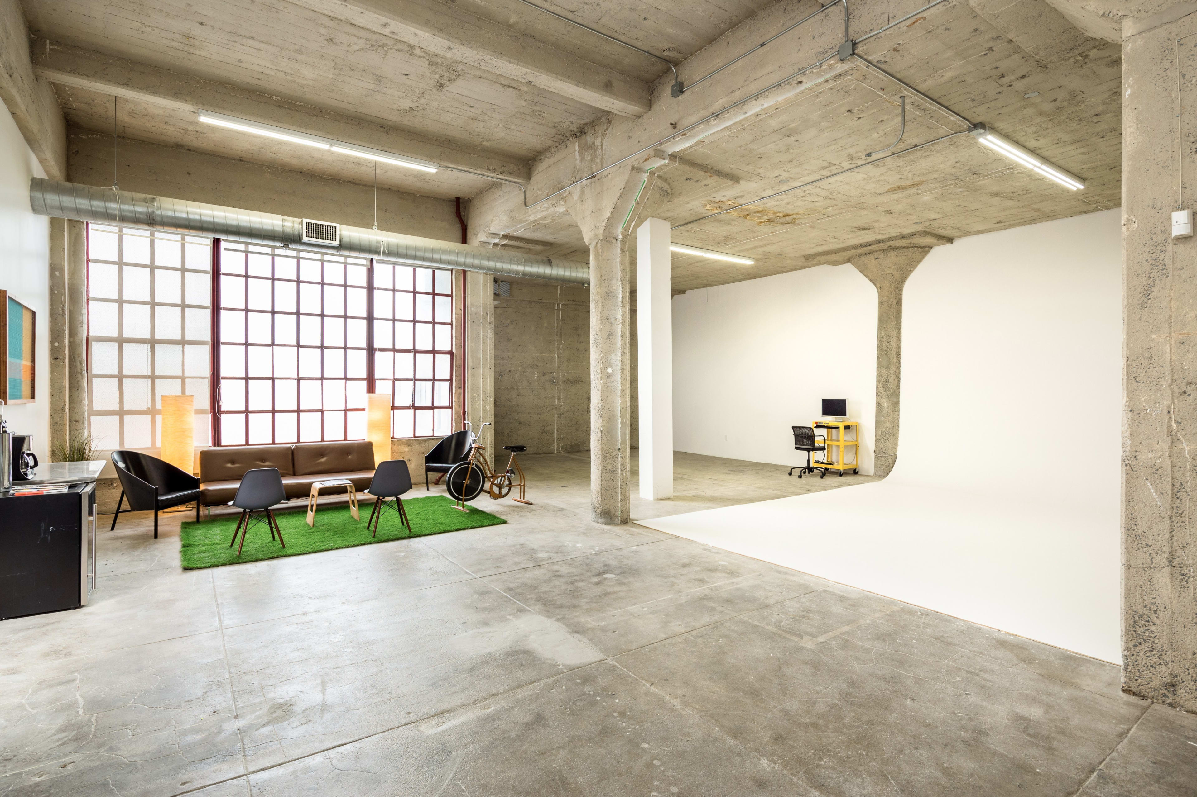 An industrial-style interior space with a seating area featuring a coffee table and green carpet, alongside a minimalistic workspace and a large window.