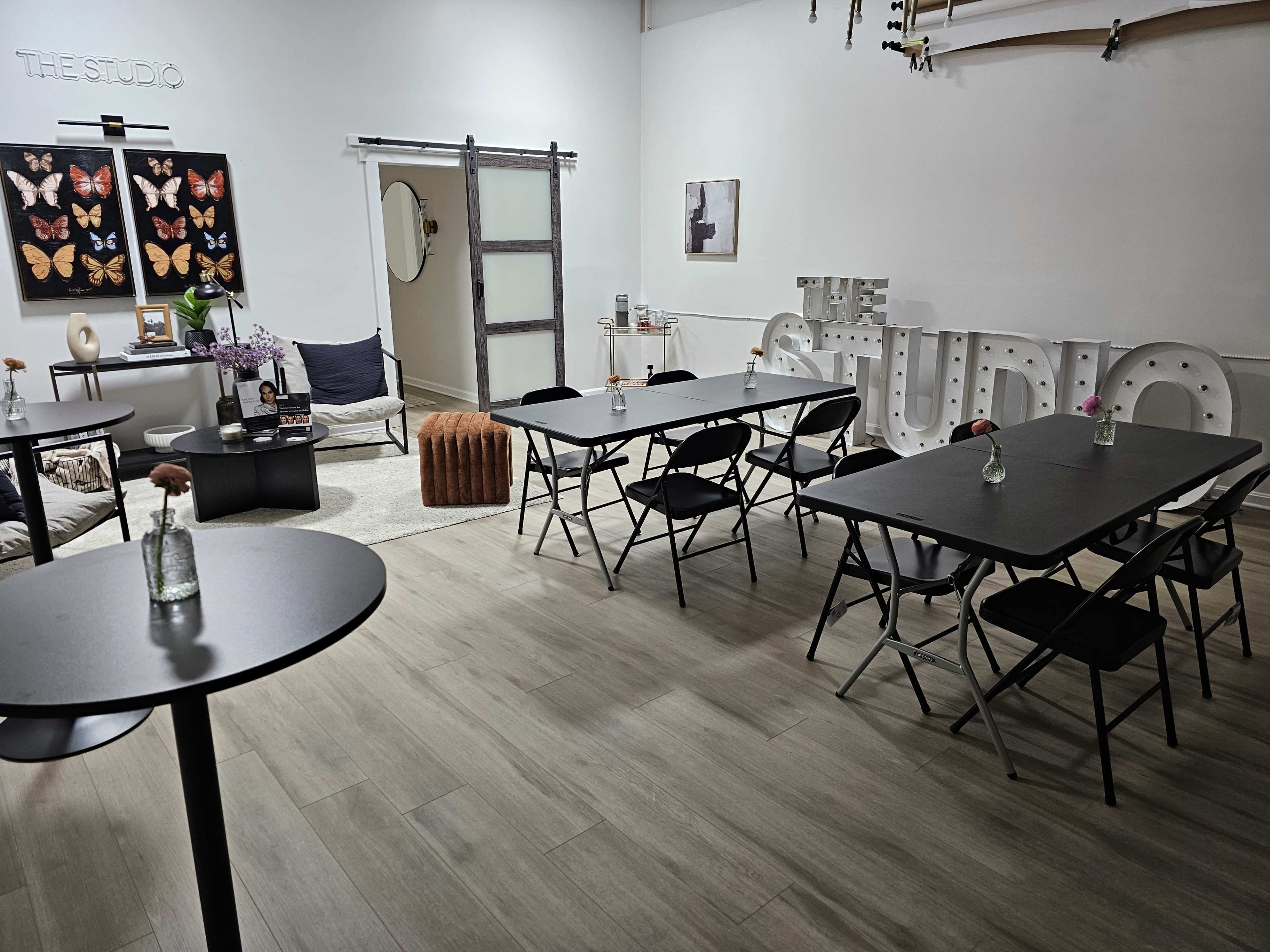 The image shows a modern studio space featuring several black tables and chairs, decorative butterfly artwork on the wall, and a cozy seating area with a large rug.