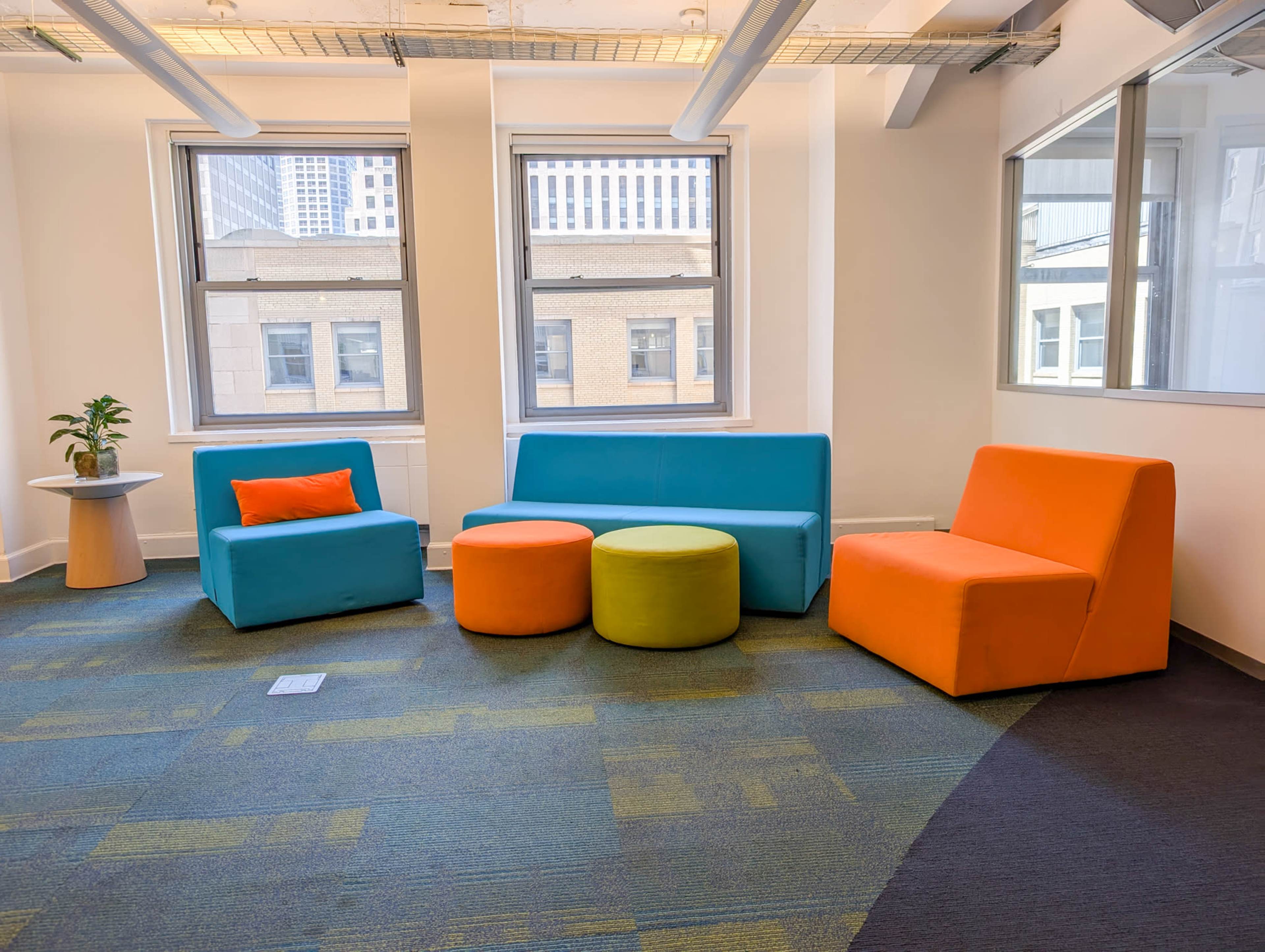 The image shows a modern seating area with brightly colored couches and poufs arranged around a small table next to large windows.