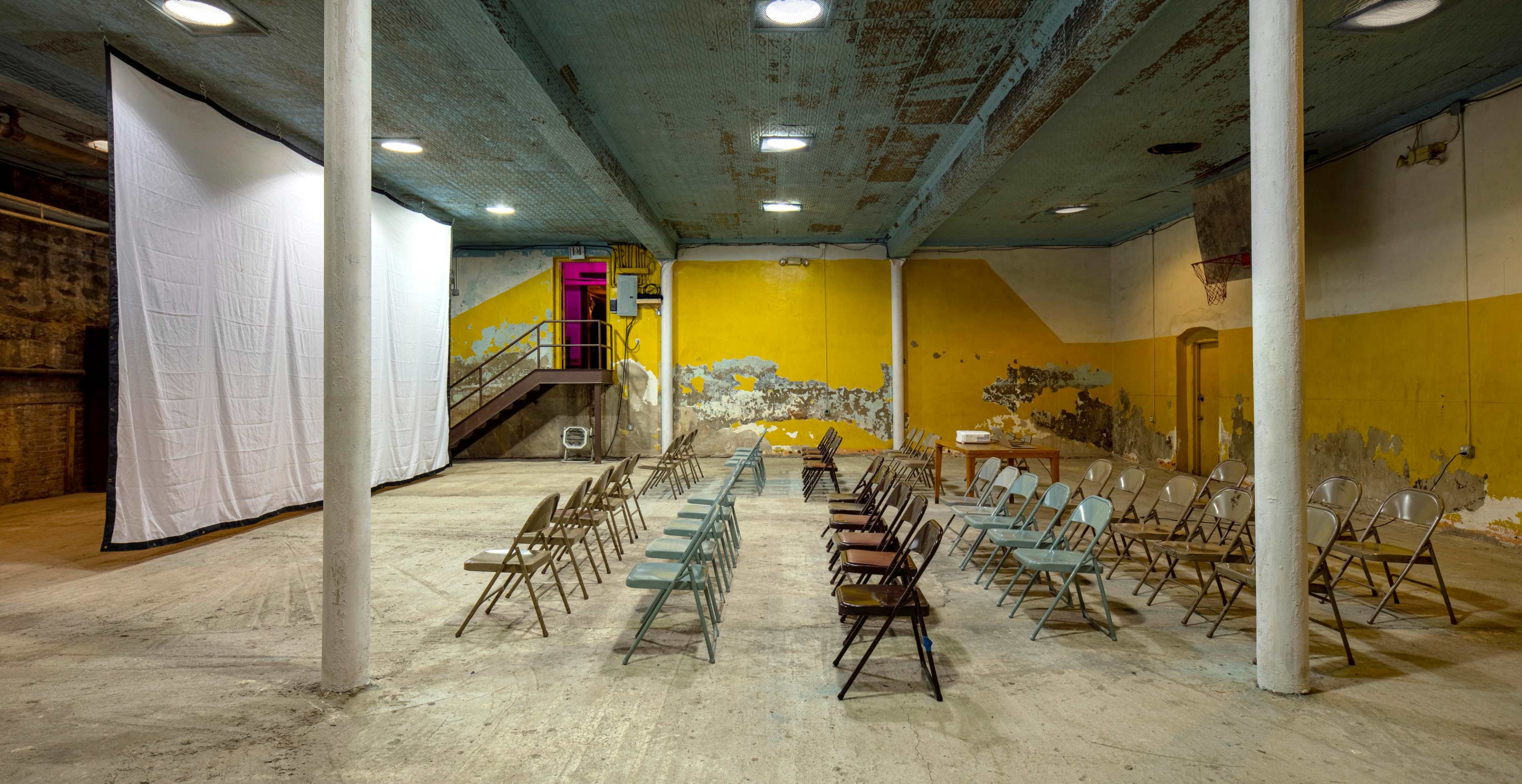 An empty industrial space features rows of folding chairs facing a large white screen, with peeling yellow and gray walls and exposed beams.