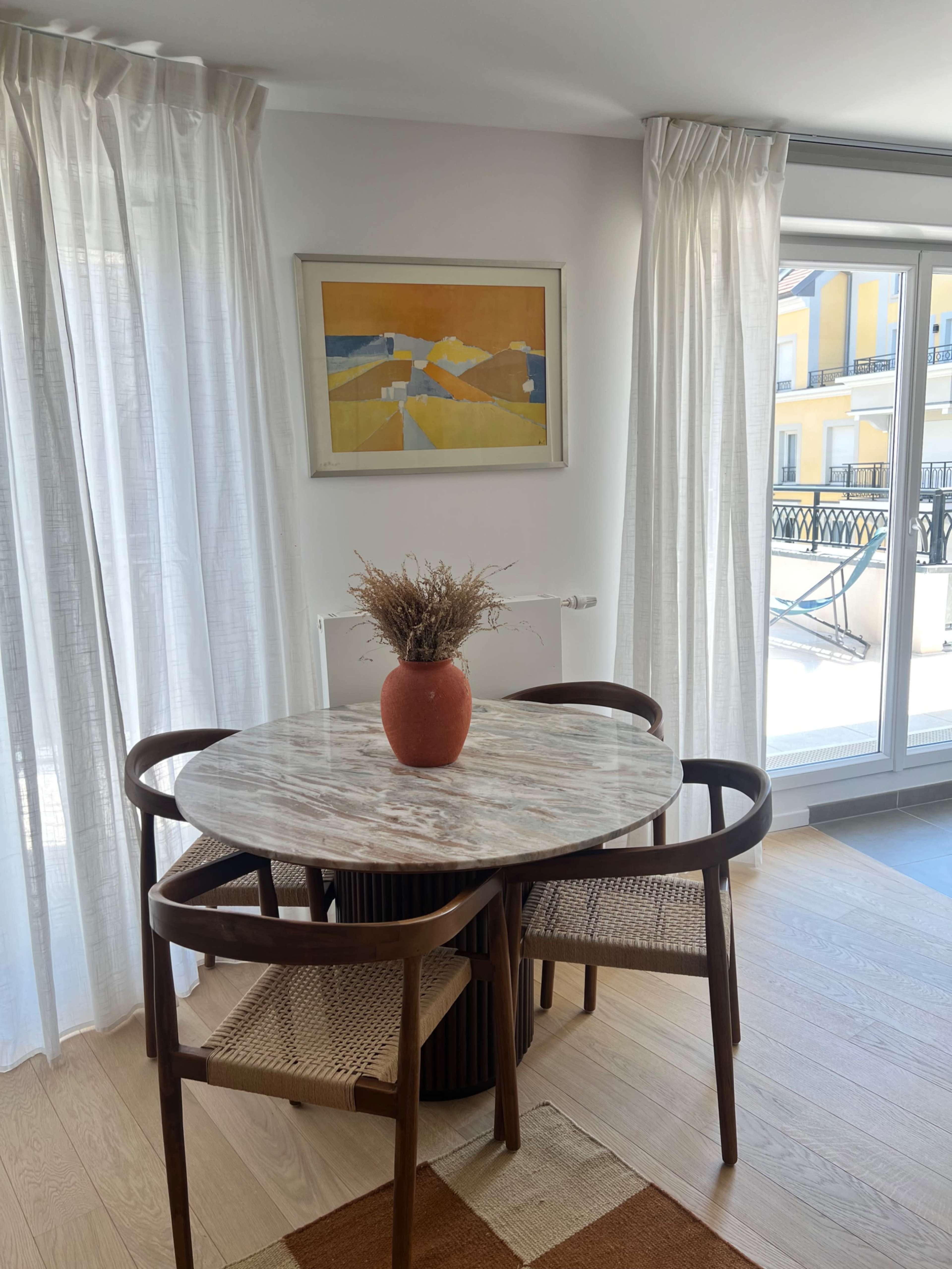 A round dining table with a marble top is placed in a brightly lit room, surrounded by four wooden chairs, and decorated with a terracotta vase holding dried flowers.