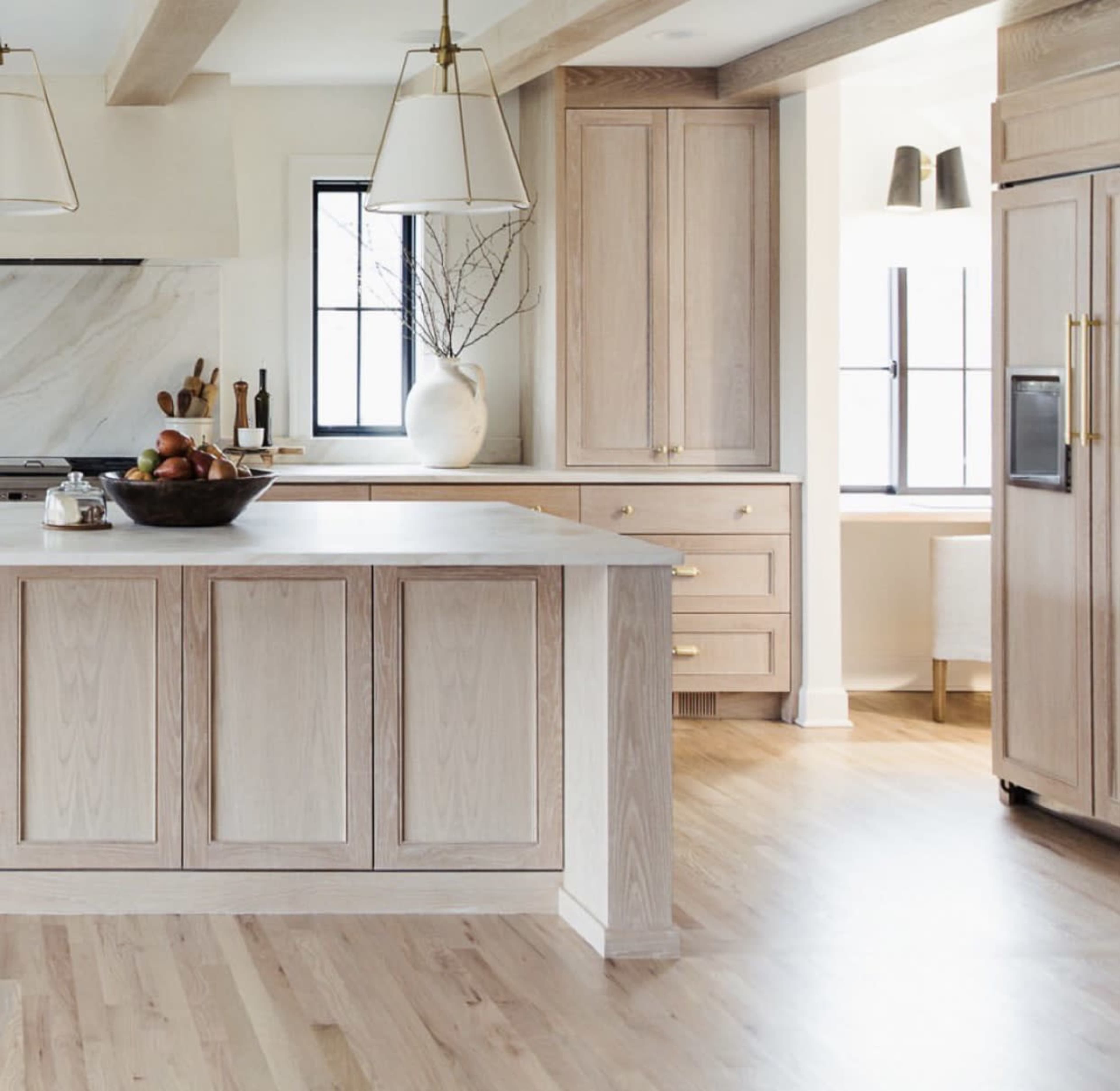 The image shows a modern kitchen featuring light wooden cabinetry, an island with a dark bowl of fruit, and ample natural light from large windows.