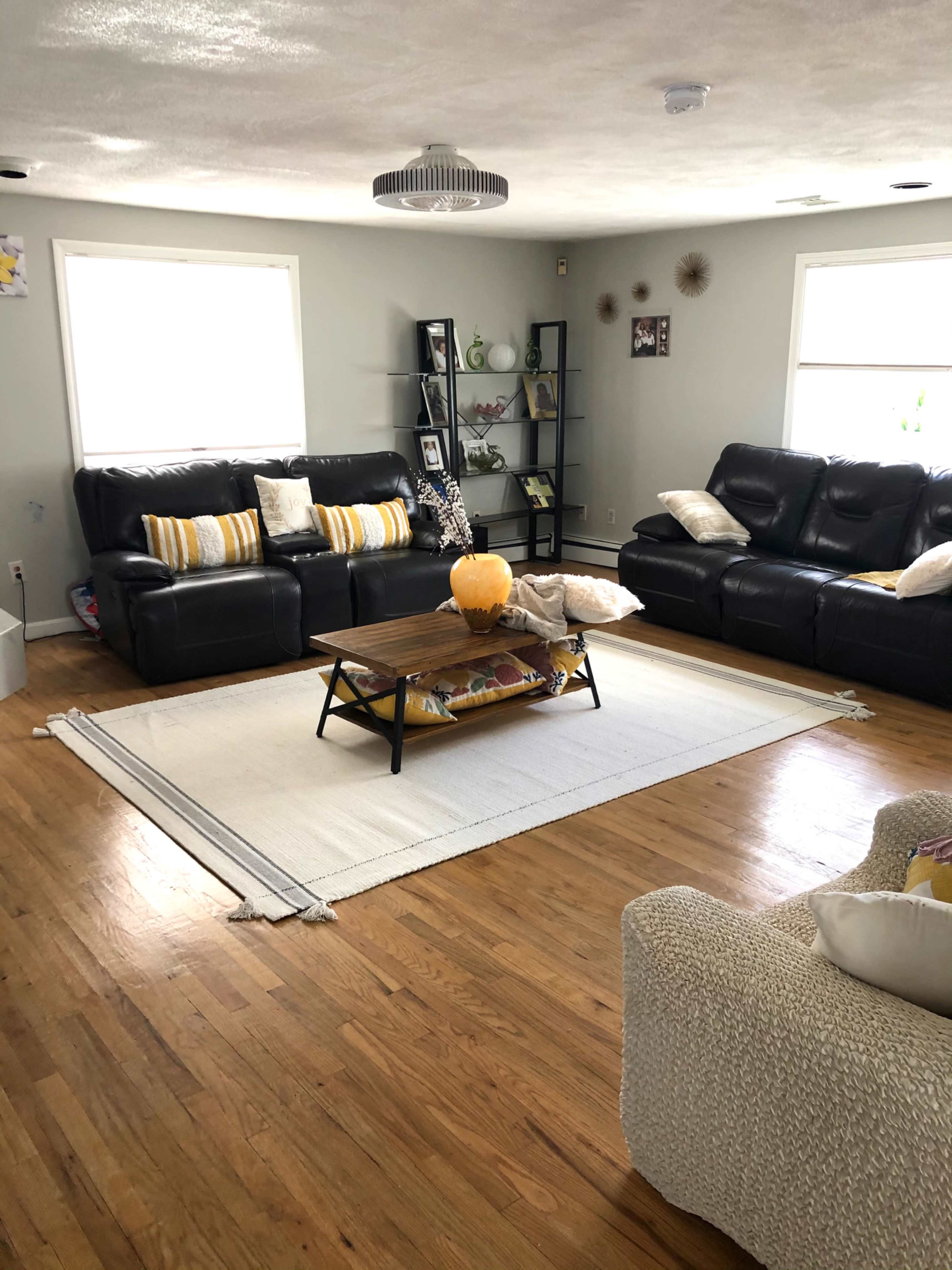 A living room with dark leather sofas, a coffee table on a rug, and a shelf displaying decorative items against light gray walls.
