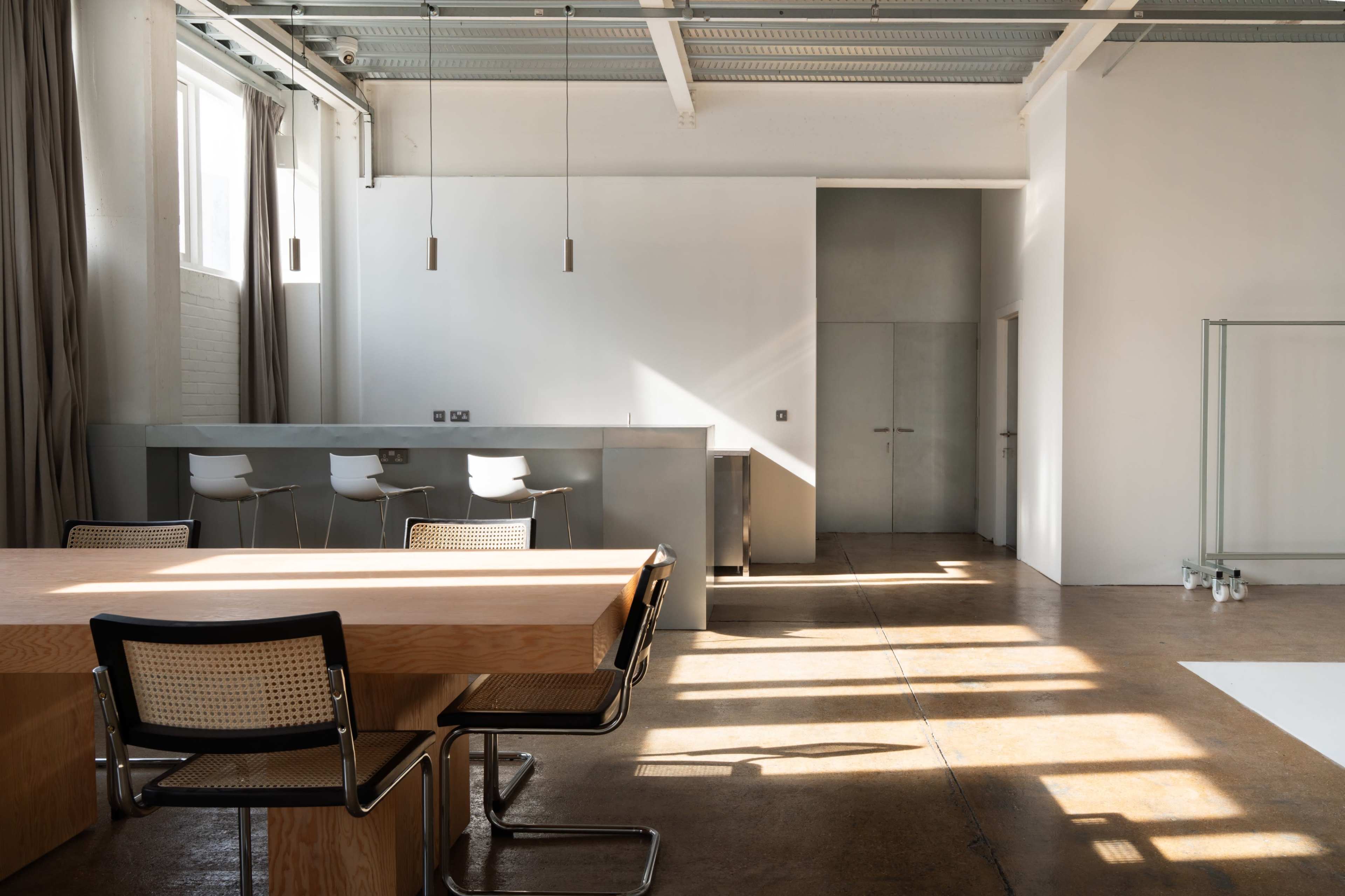 The image shows a spacious, modern interior with a large wooden table surrounded by chairs, a minimalist kitchenette, and natural light streaming through large windows.