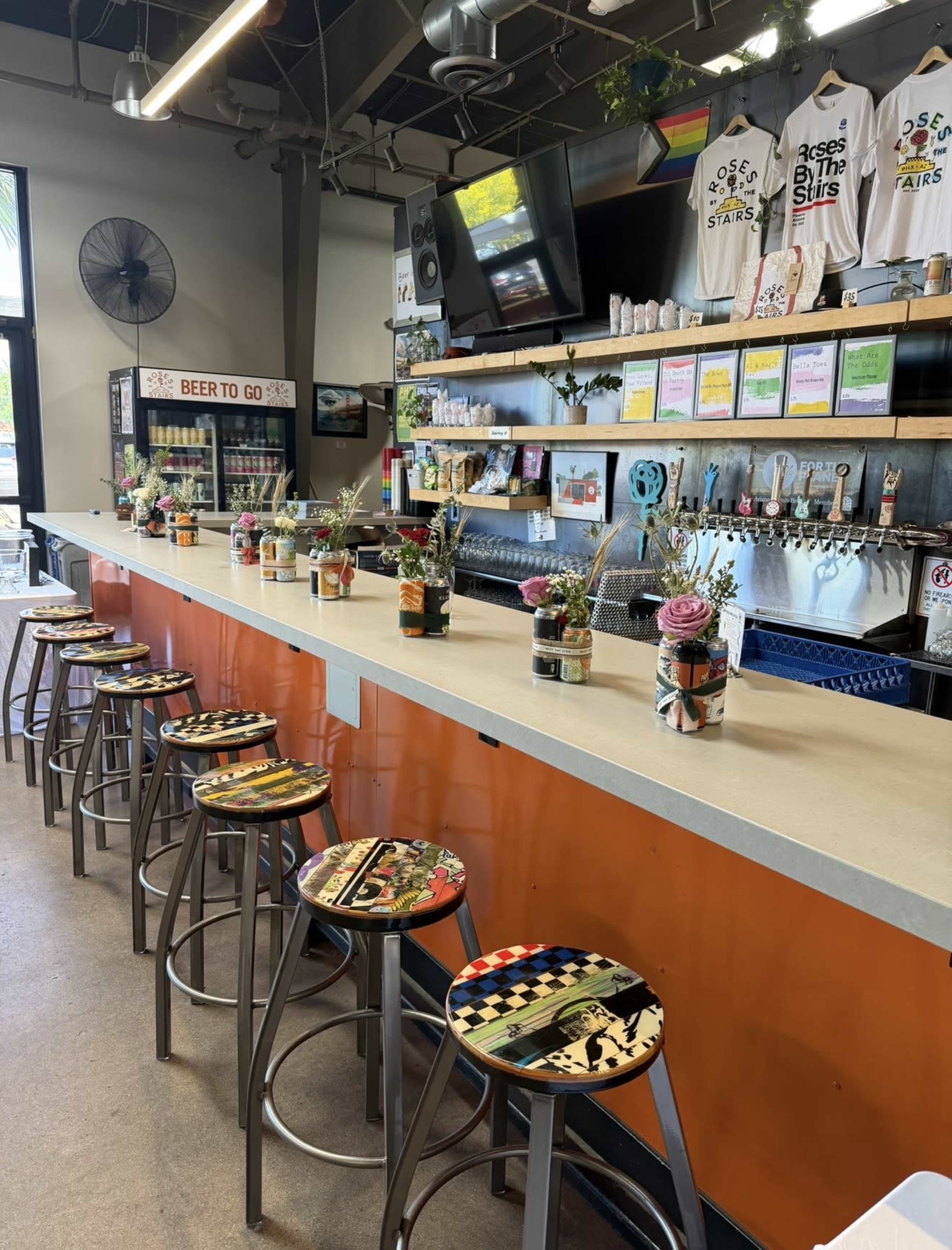 The image shows a brightly colored bar area with metal stools, decorative flowers in jars on the counter, and a variety of posters and menus displayed on the walls.