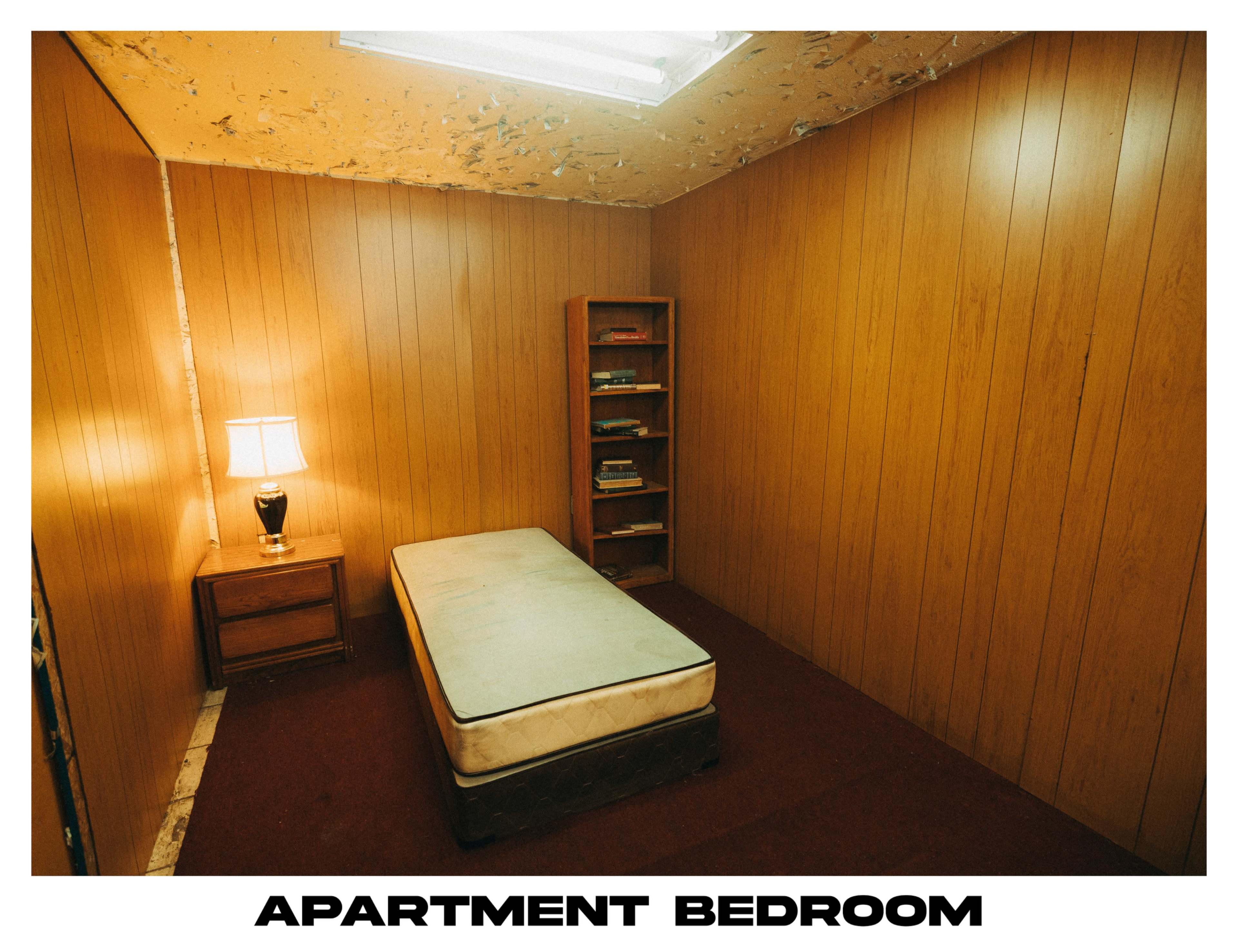 A small, sparsely furnished bedroom with wood-paneled walls, a mattress on a bed frame, a nightstand with a lamp, and a bookshelf.