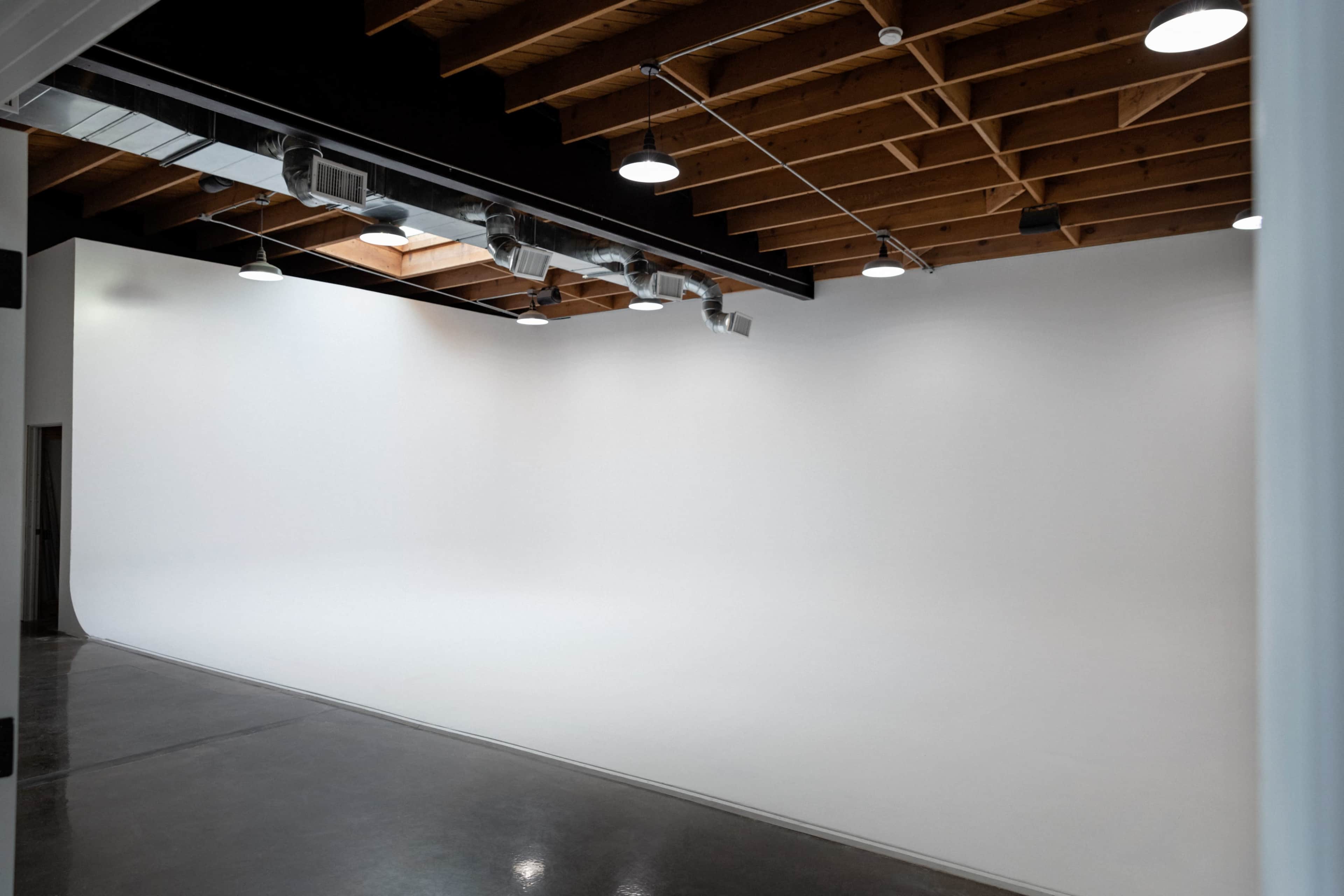 The image shows a spacious, empty studio with a seamless white backdrop, wooden beams, and bright overhead lighting.