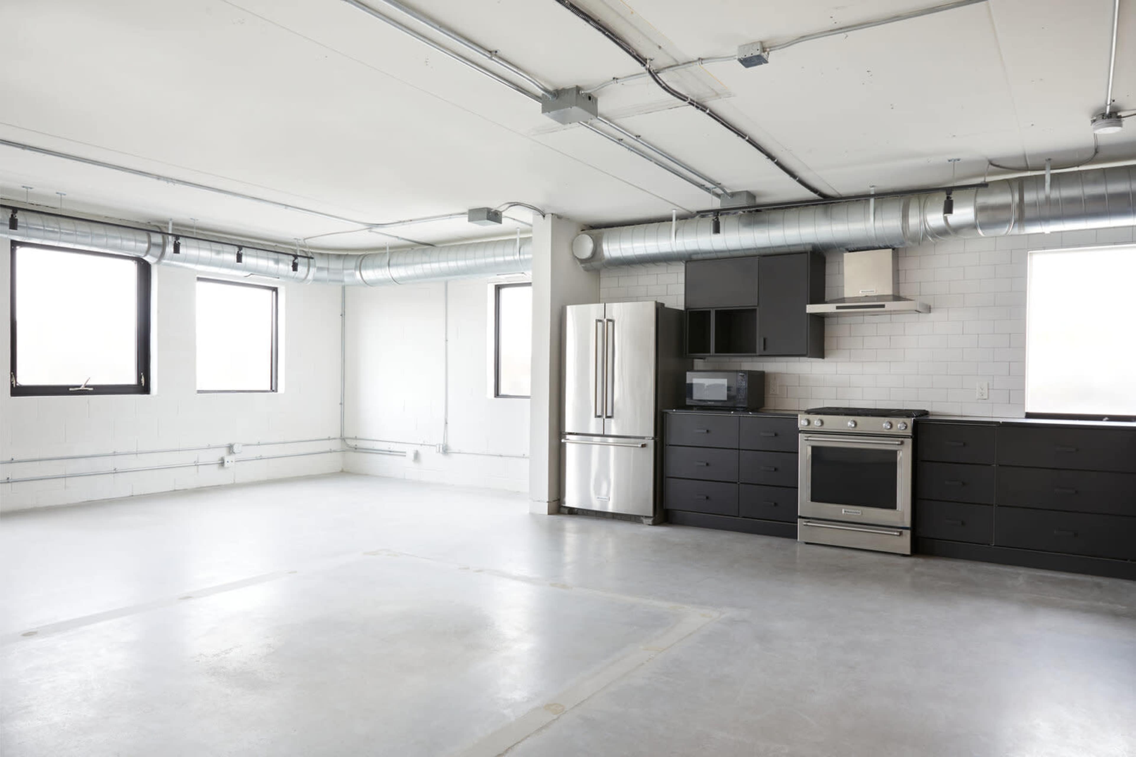 The image shows a spacious, modern kitchen area with stainless steel appliances and concrete flooring, featuring large windows on two walls.