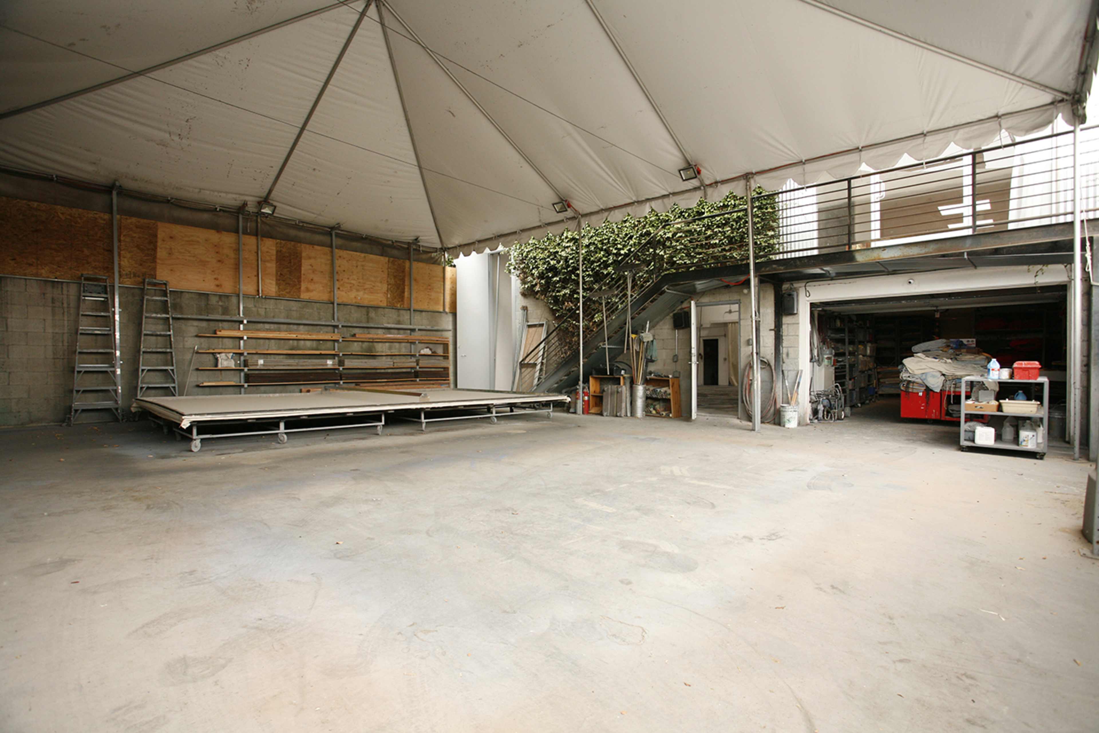 The image shows a large, empty tent structure with a wooden platform on one side and storage areas visible in the background.