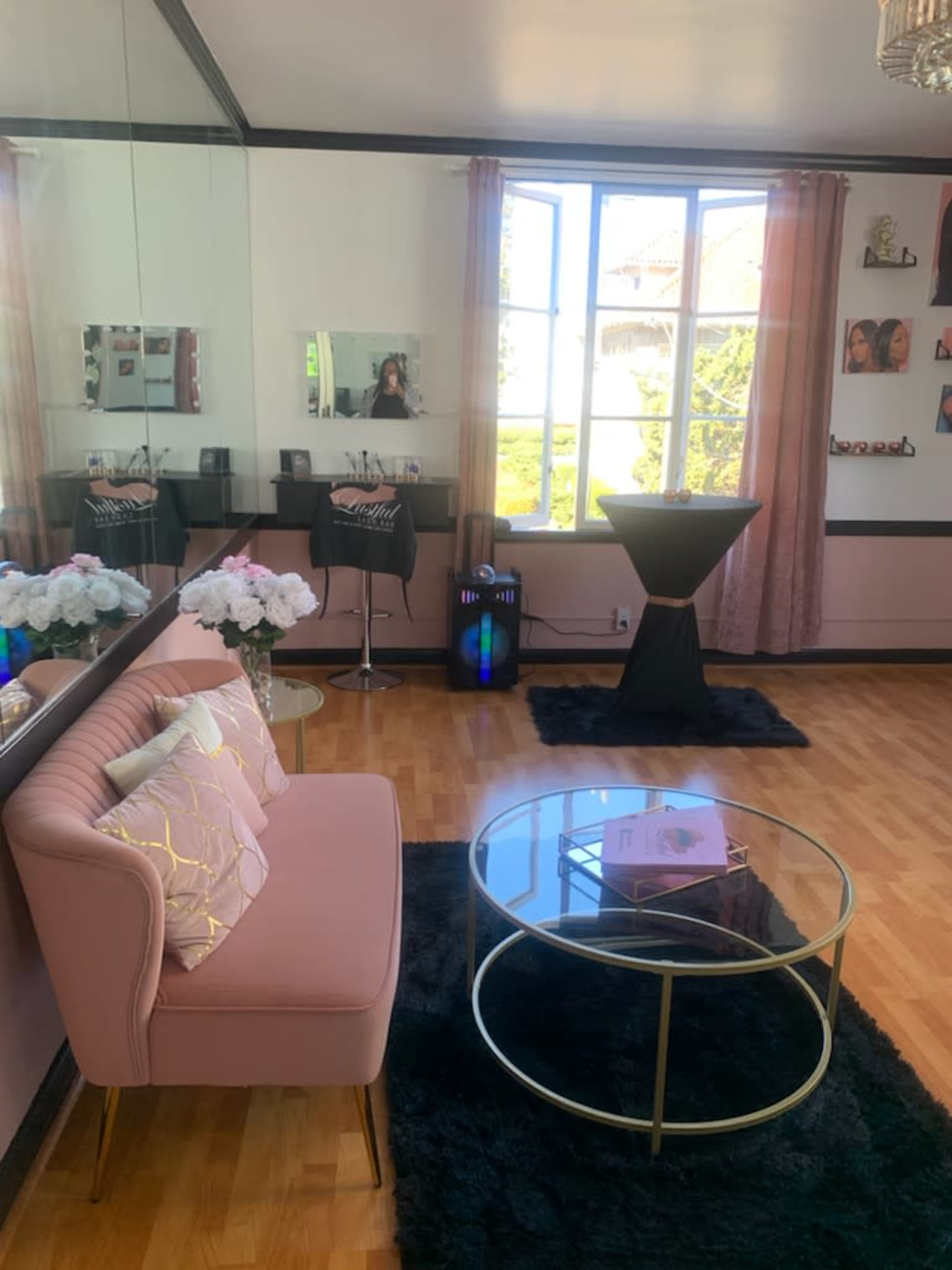 The image shows a well-decorated room featuring a pink sofa, a glass coffee table, and a tall black table near large windows with floral arrangements.