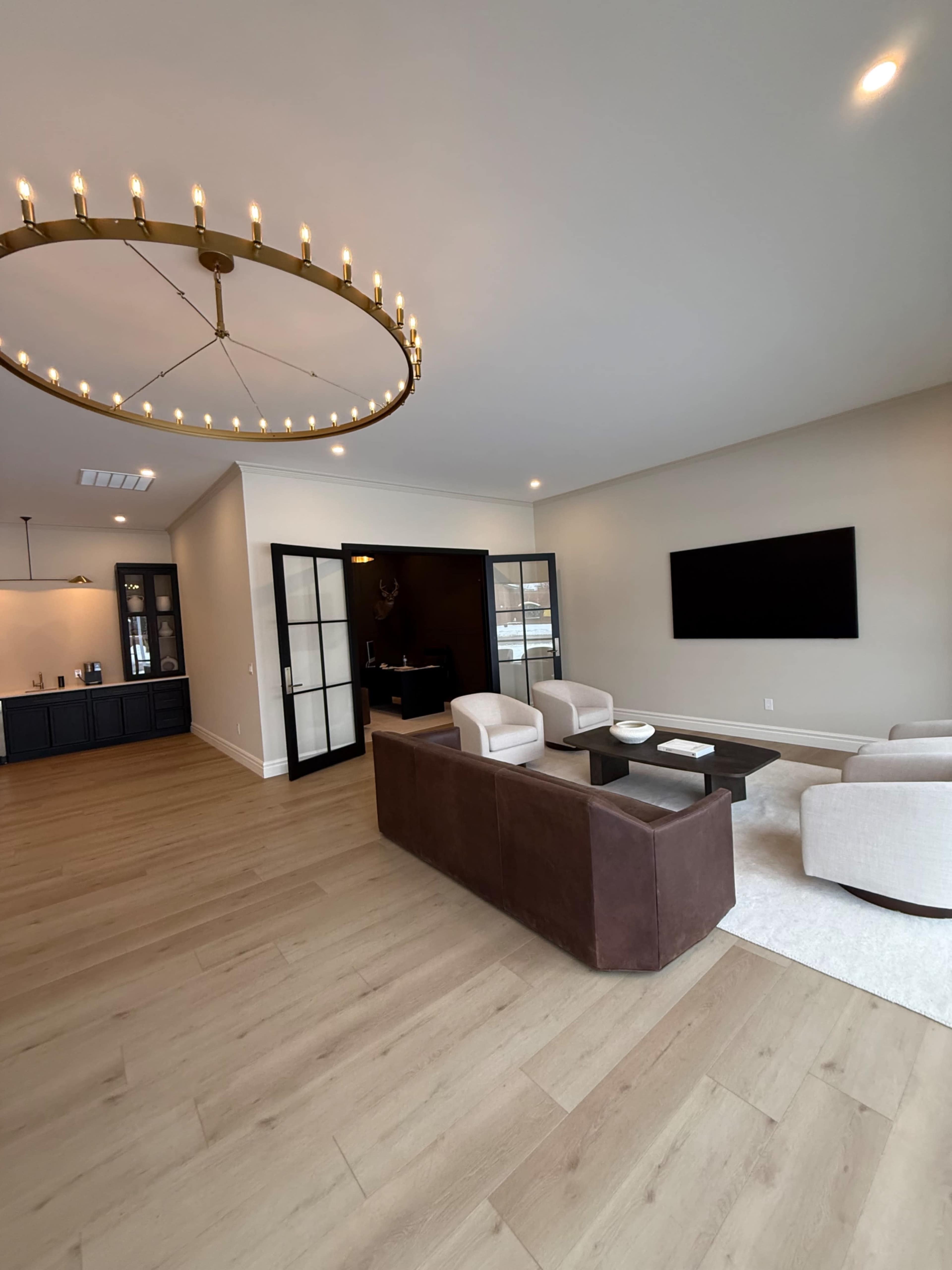 The image shows a spacious, modern living room featuring a circular chandelier, a dark wood sideboard, a large TV mounted on the wall, and a sectional sofa with two light-colored armchairs.