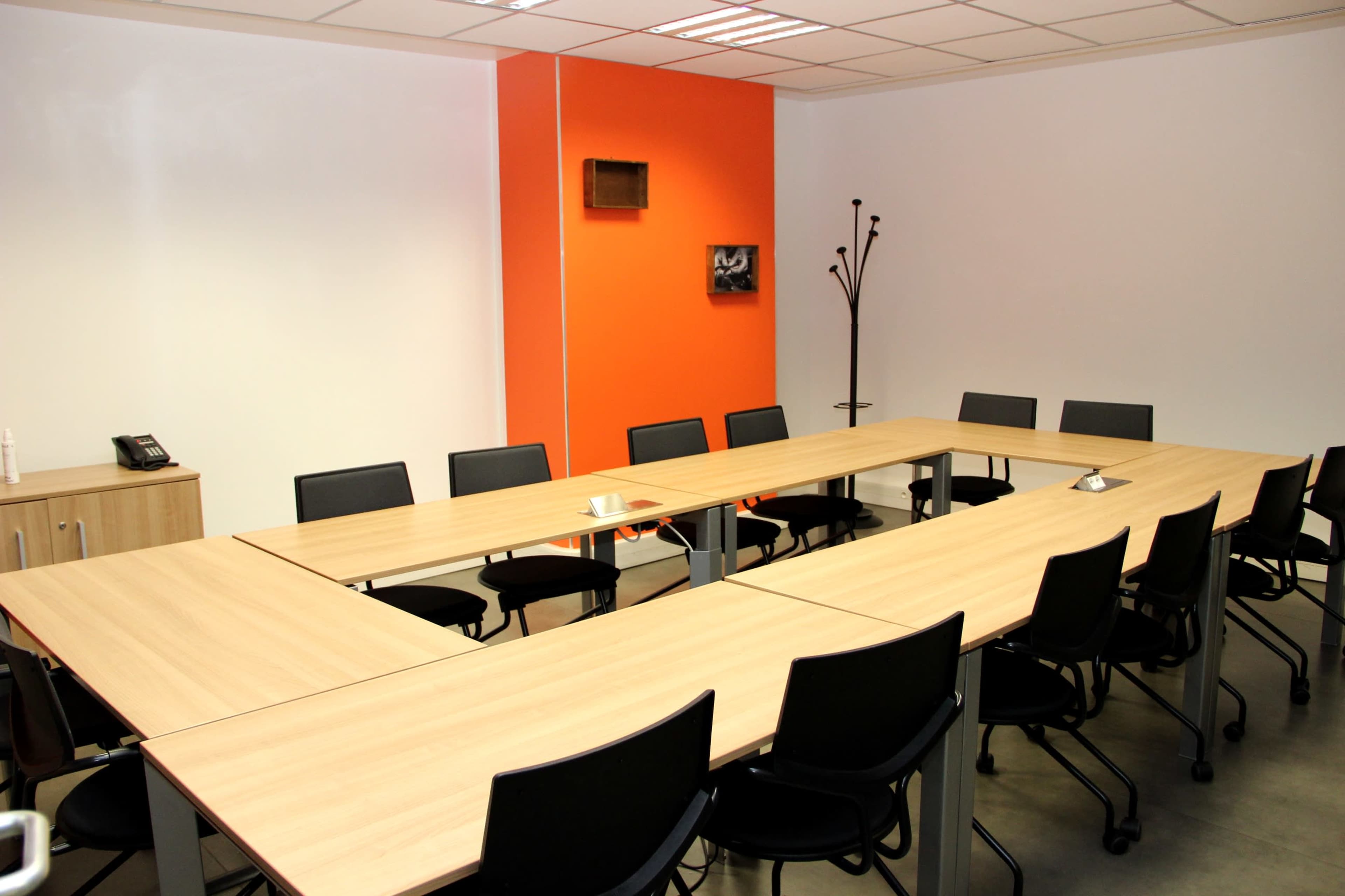 A conference room with a rectangular table arrangement surrounded by black chairs, featuring an orange accent wall and minimal decor.