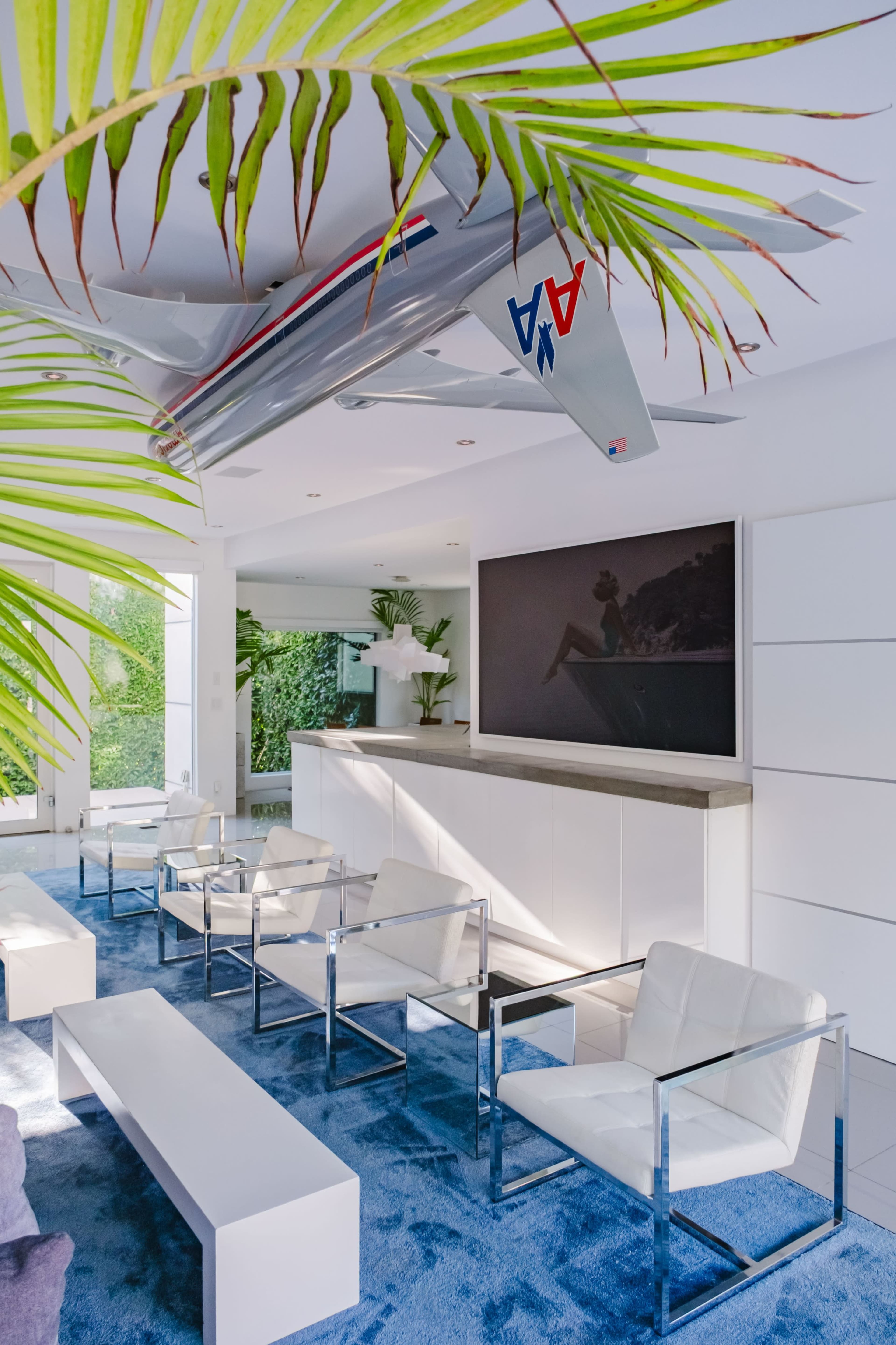 A modern living room features white furniture and a large television mounted above a sleek cabinet, with an airplane sculpture suspended from the ceiling and greenery in the background.