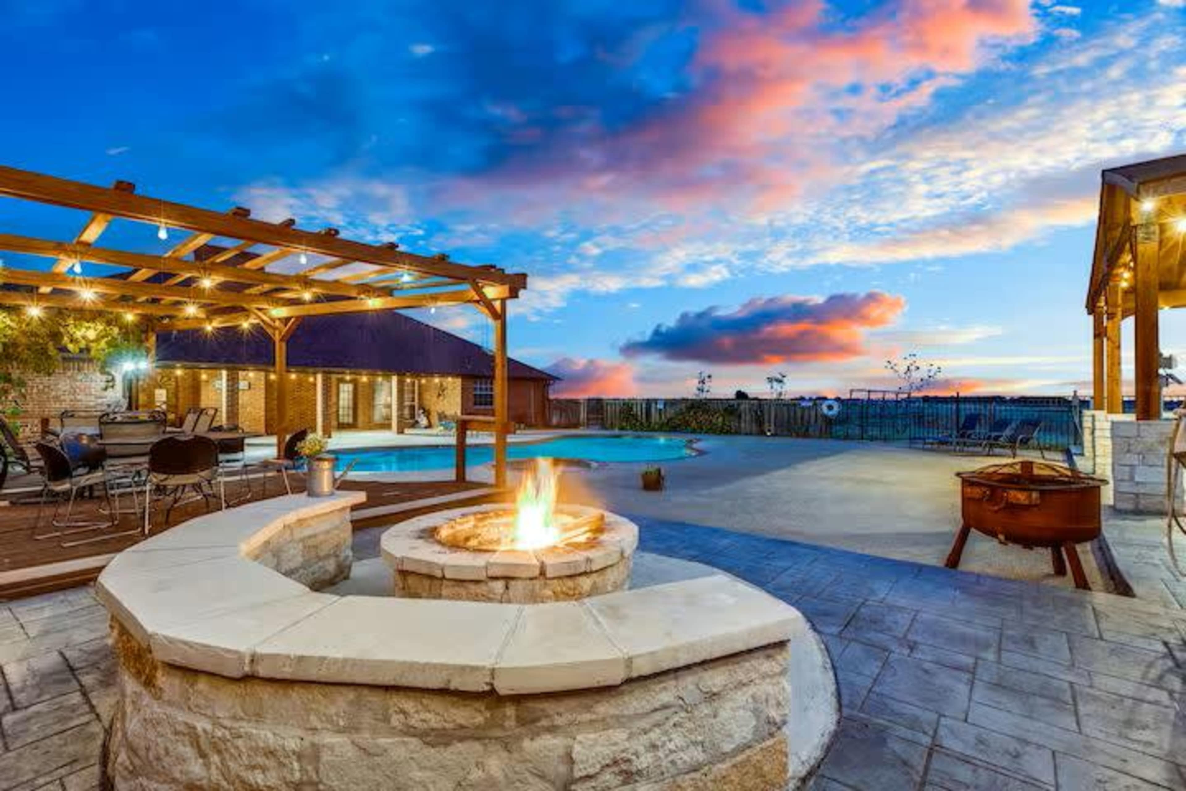 A backyard setting with a stone fire pit, a swimming pool nearby, and a covered patio area under a colorful sunset.