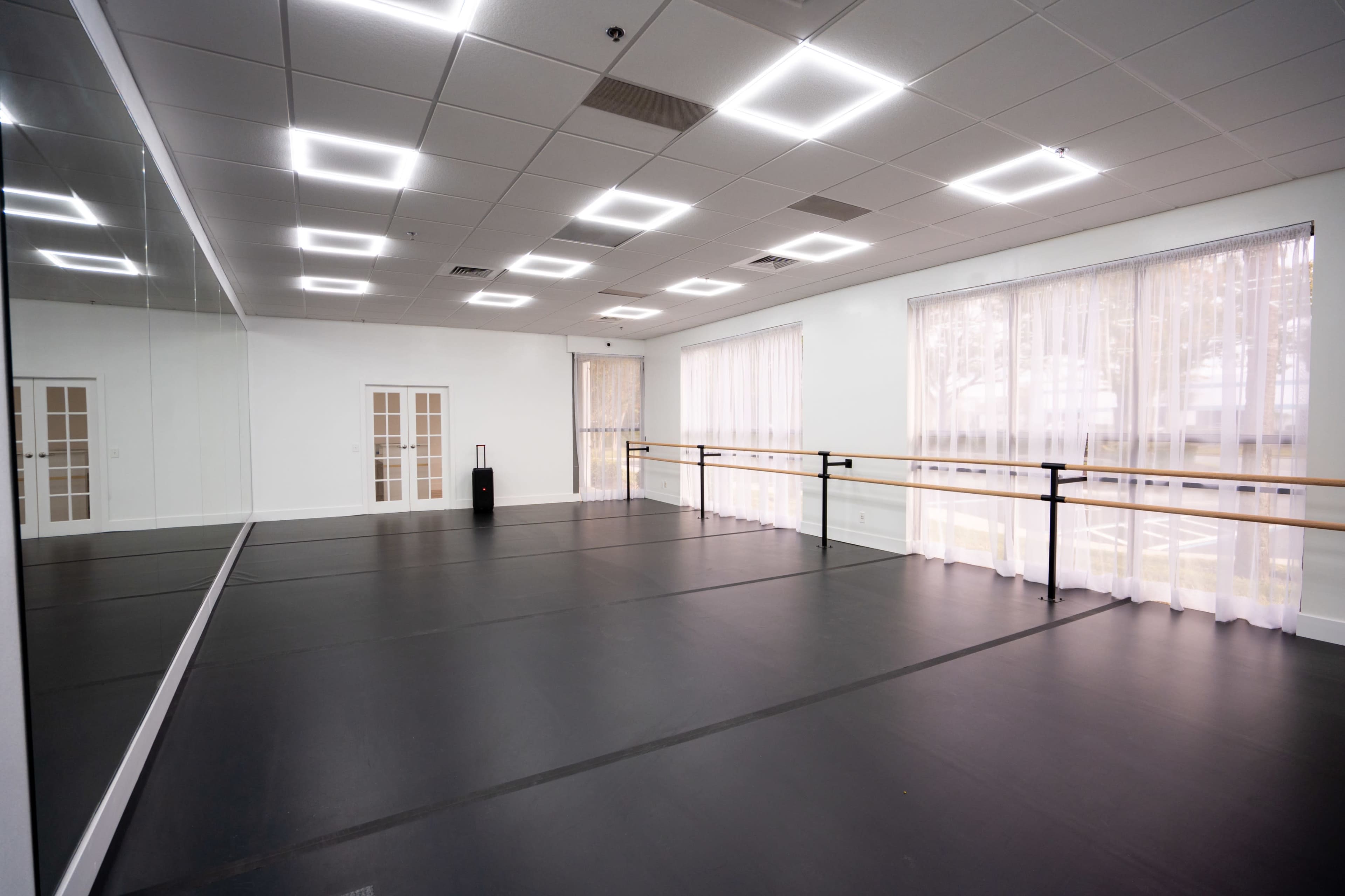 The image shows an empty dance studio with a mirror along one wall, wooden ballet bars, and large windows covered with sheer curtains.