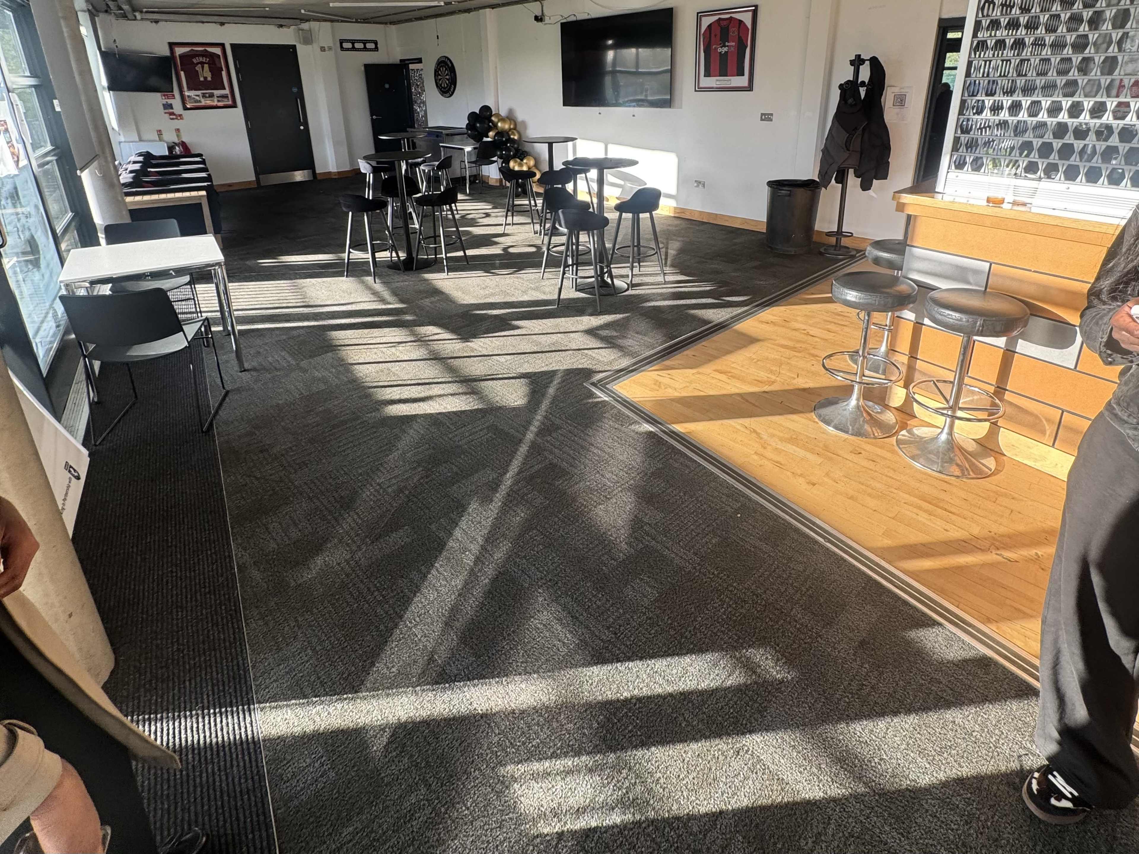 The image shows an interior space with tables and chairs, hardwood flooring, and large windows allowing sunlight to cast shadows across the carpeted area.