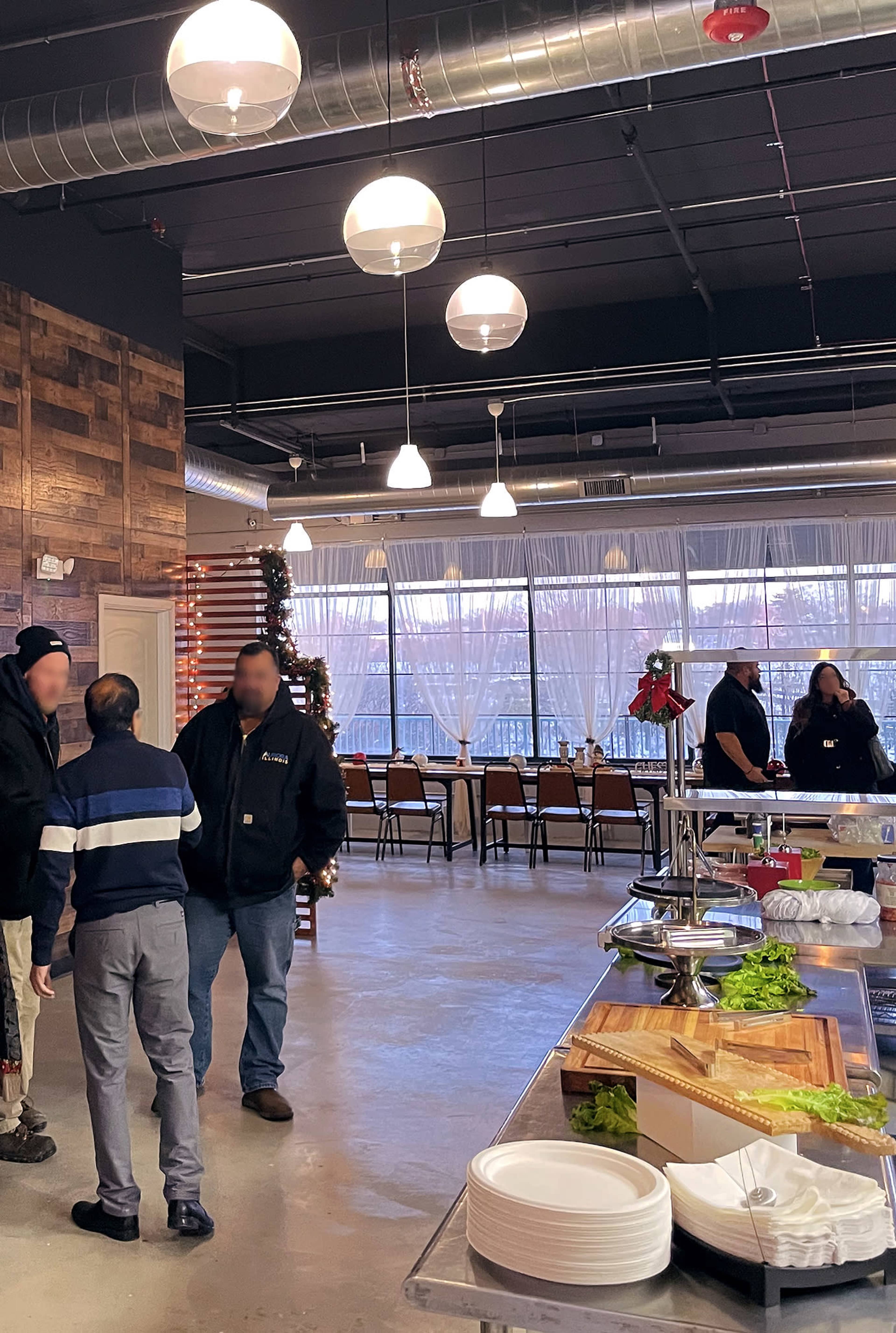 The image shows a modern event space with a buffet setup, featuring tables and chairs arranged in the background and several individuals gathered in conversation.